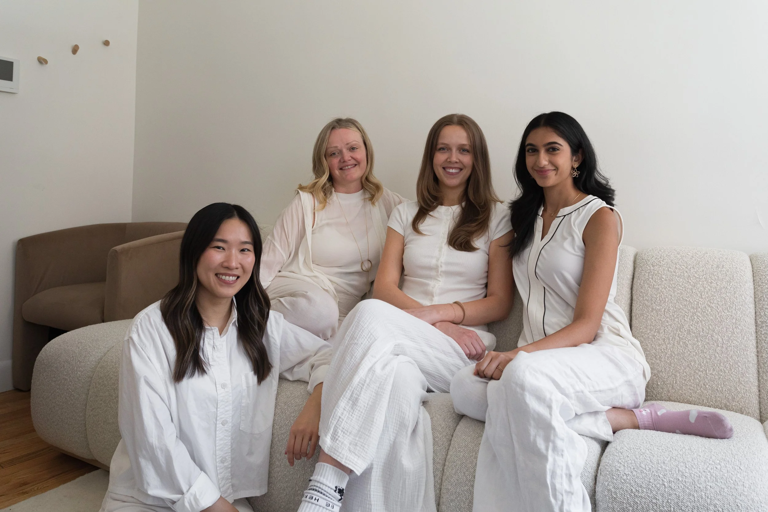 A group of five women sitting on a beige couch in a minimalistic room, smiling at the camera. They are dressed in white casual clothing.