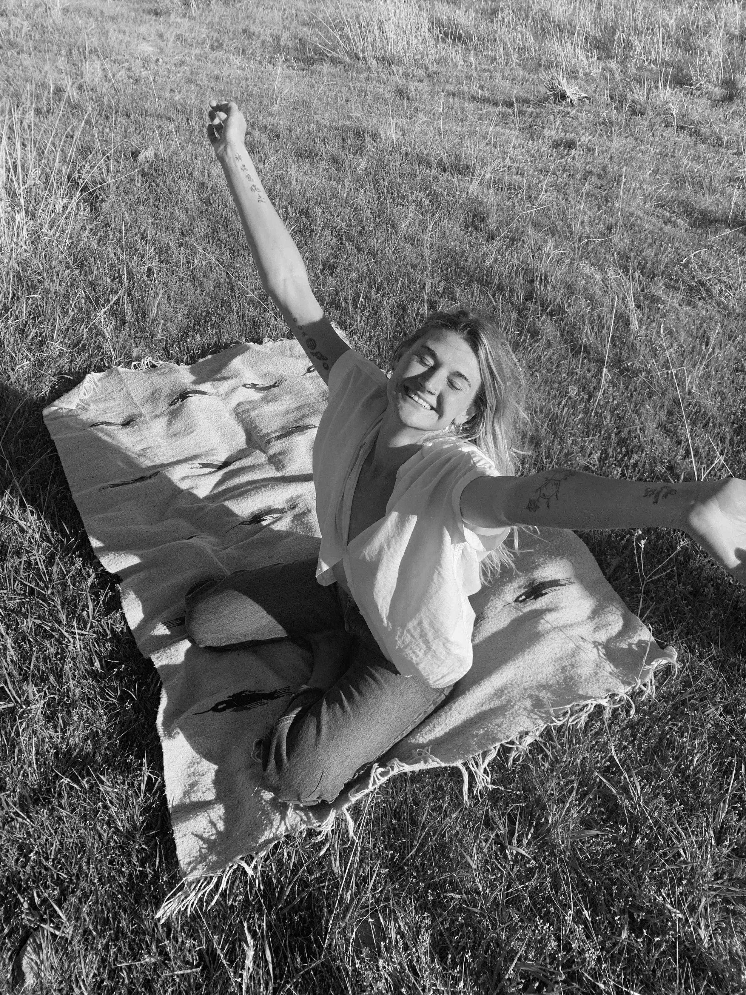 A young woman with blonde hair, sitting on a blanket in a grassy field, smiling with eyes closed and arms outstretched to the sides.