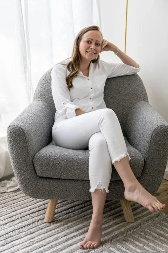 Woman sitting in a gray armchair, smiling, with her legs crossed at the ankles, wearing a white shirt and white jeans, in a bright room with white curtains and a striped rug.