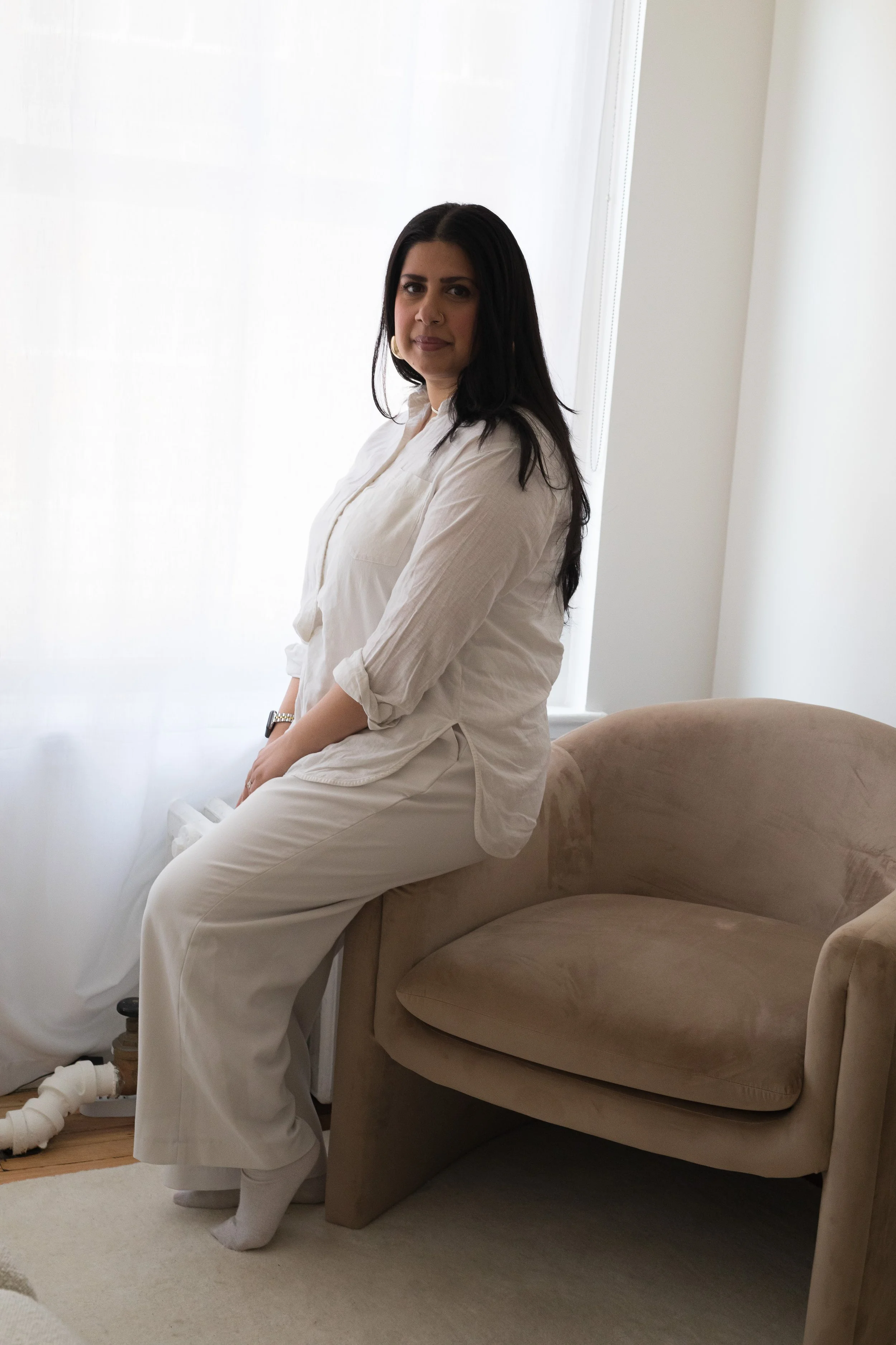 A woman with long dark hair sitting on a beige curved couch in a room with white walls and a window with white curtains.