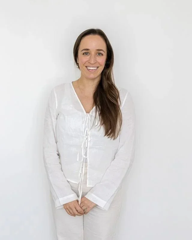 A woman with long brown hair, smiling, wearing a white blouse with a tie-front detail and white pants, standing against a plain white wall.