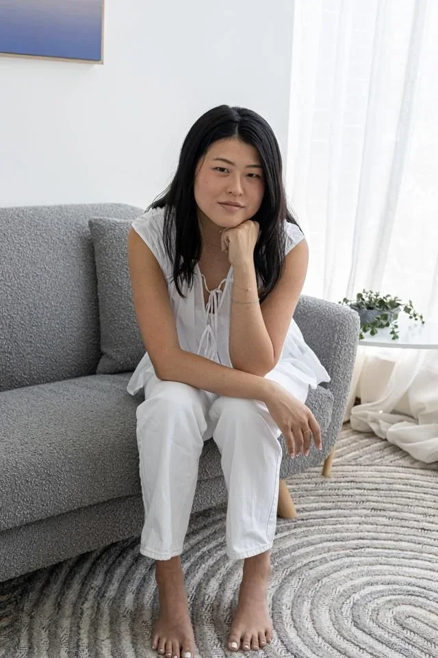 A woman sitting on a gray sofa in a bright room with white walls and curtains, wearing a white sleeveless top and white pants, resting her chin on her hand and looking at the camera.