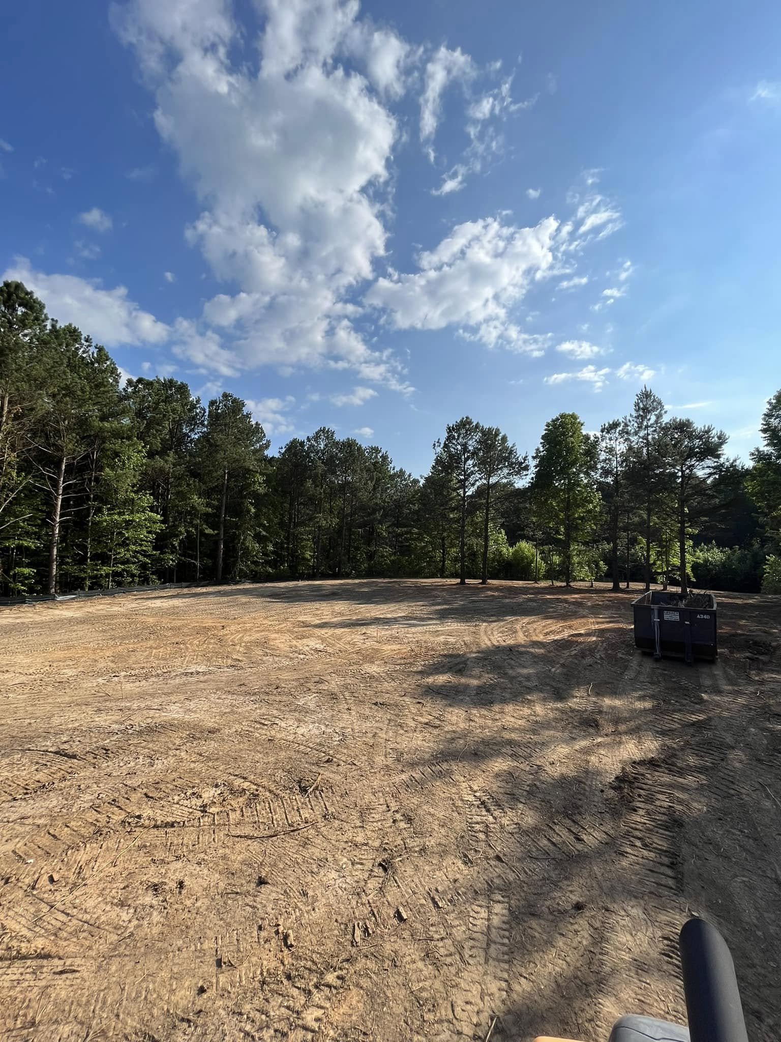 Clear sky with scattered clouds above a cleared dirt area bordered by tall green trees; a black container is visible on the right side of the image.