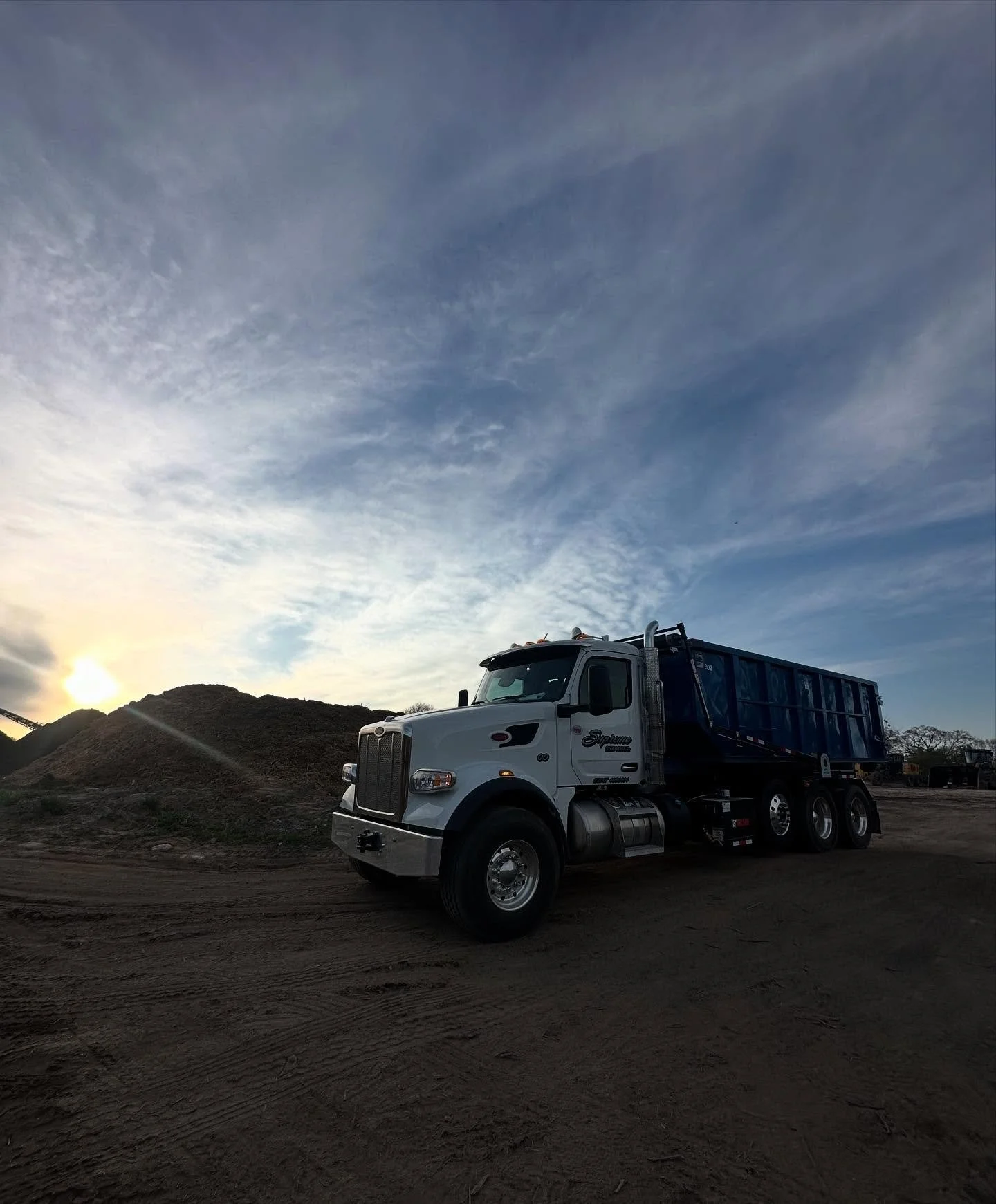 A white semi-truck with a blue trailer parked on a dirt lot with mounds of dirt behind it, under a sky with scattered clouds during sunset.
