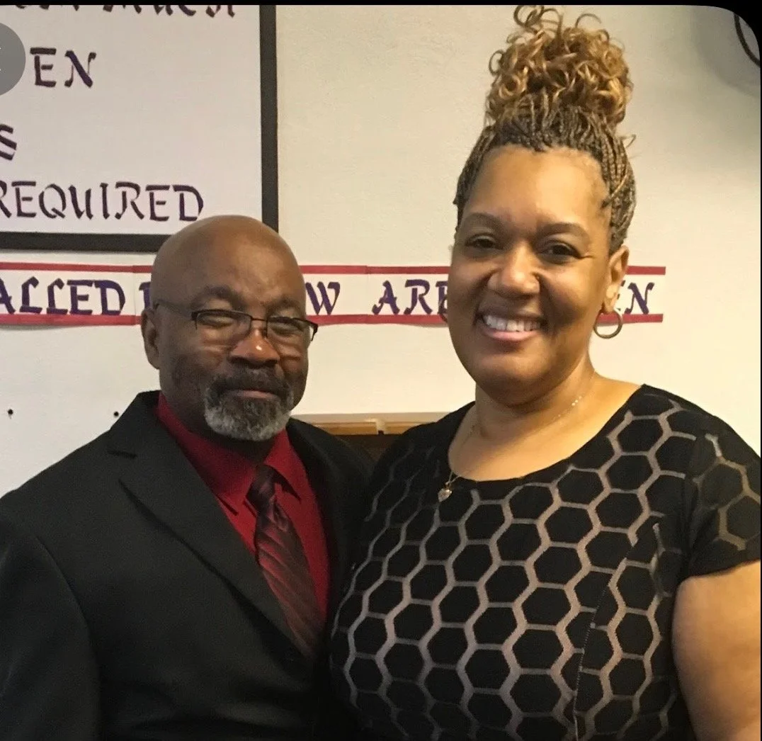 A man and woman smiling and standing close together indoors with a sign behind them.