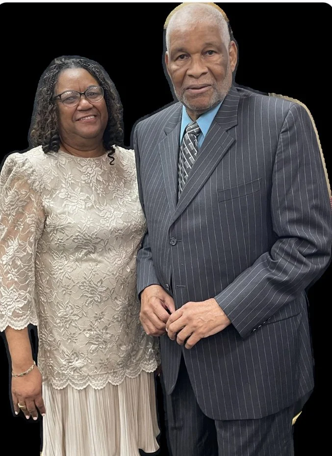 An elderly man in a pinstripe suit and a woman in a cream lace dress standing together against a black background.