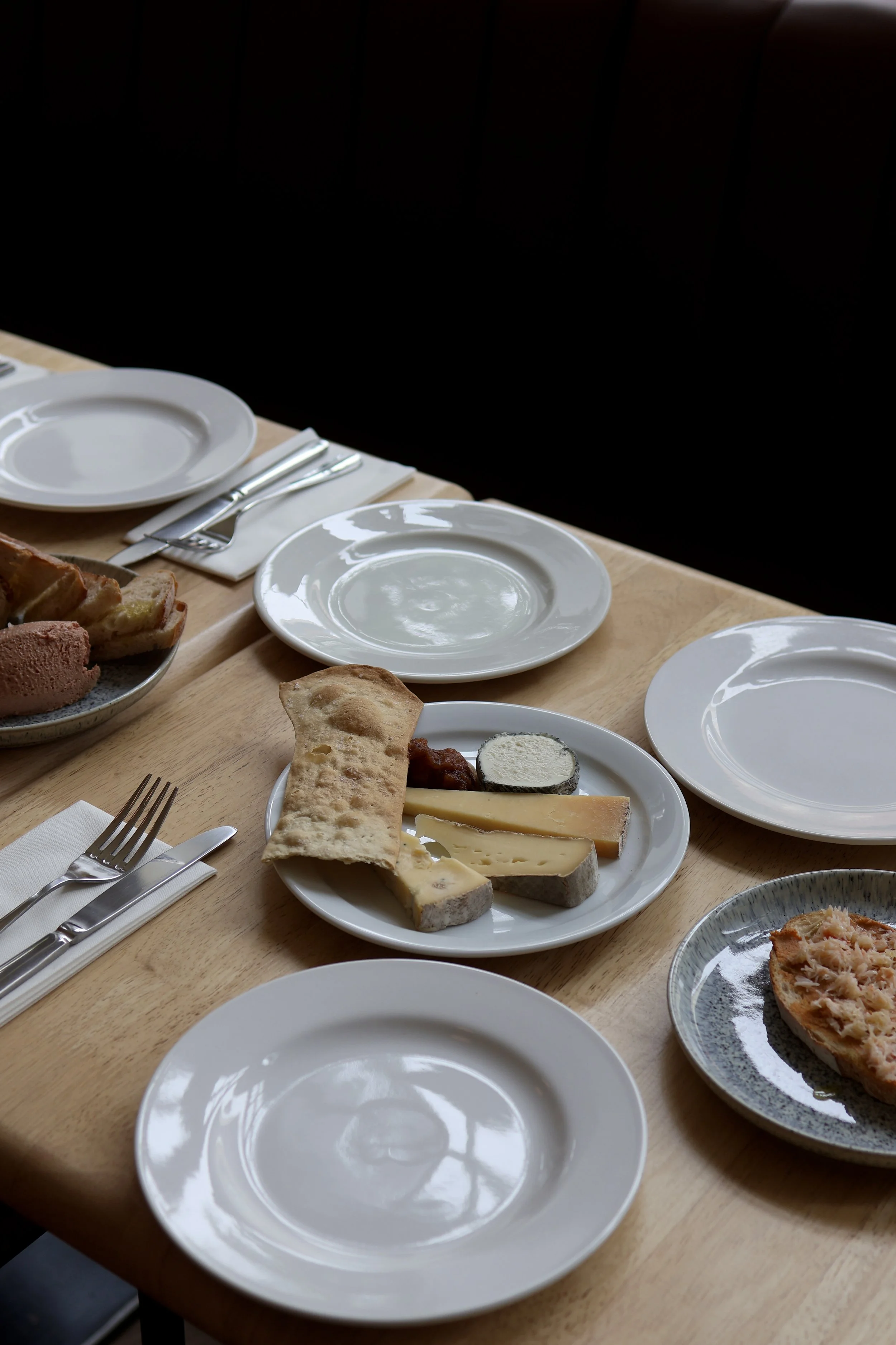 A table set for a meal with empty white plates, silverware, and a cheese platter with various cheeses, bread, and a spread. The table is wooden, and the background is dark.