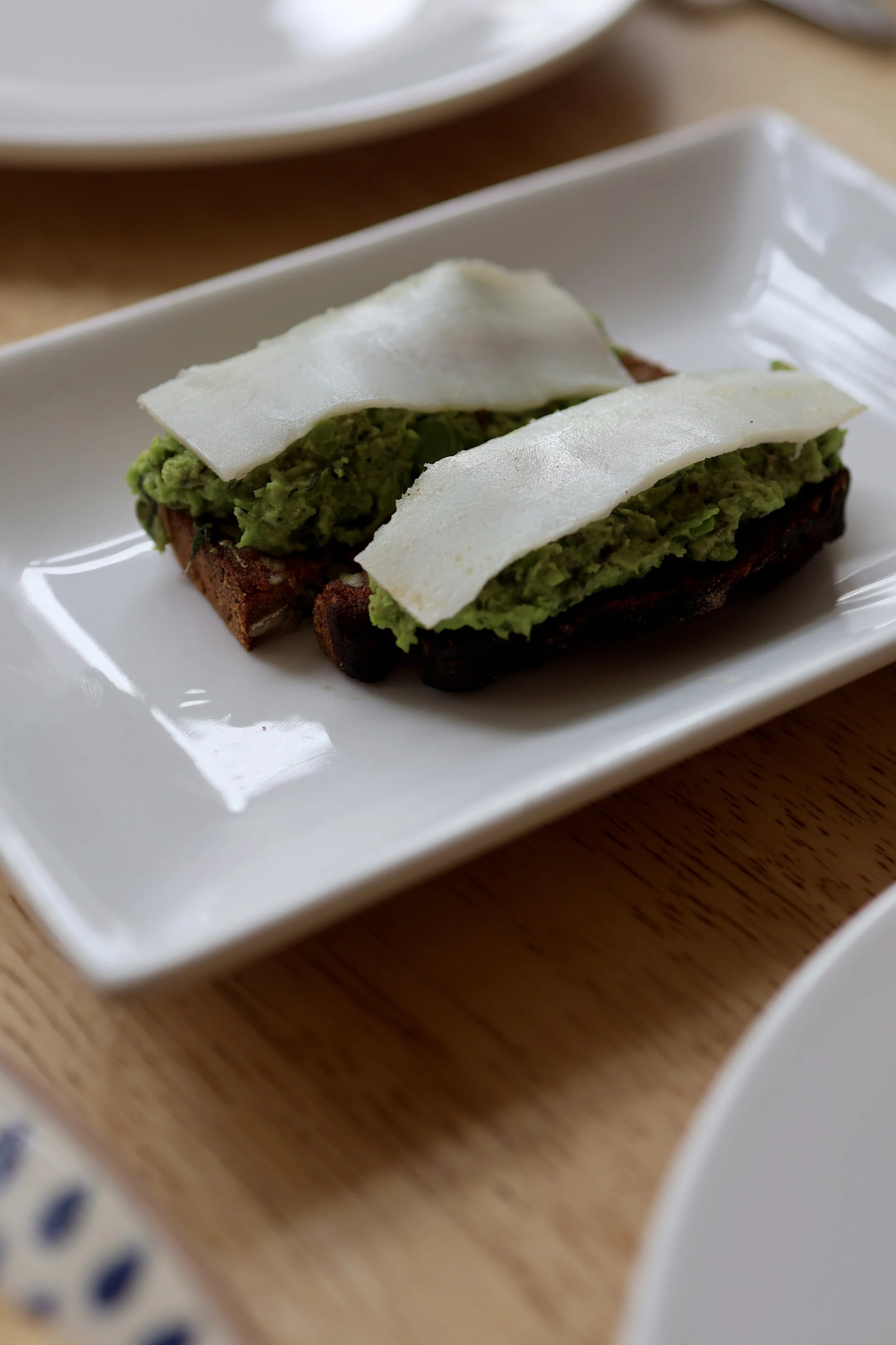 Two pieces of toasted bread topped with smashed avocado and thin slices of cheese, served on a white rectangular plate.