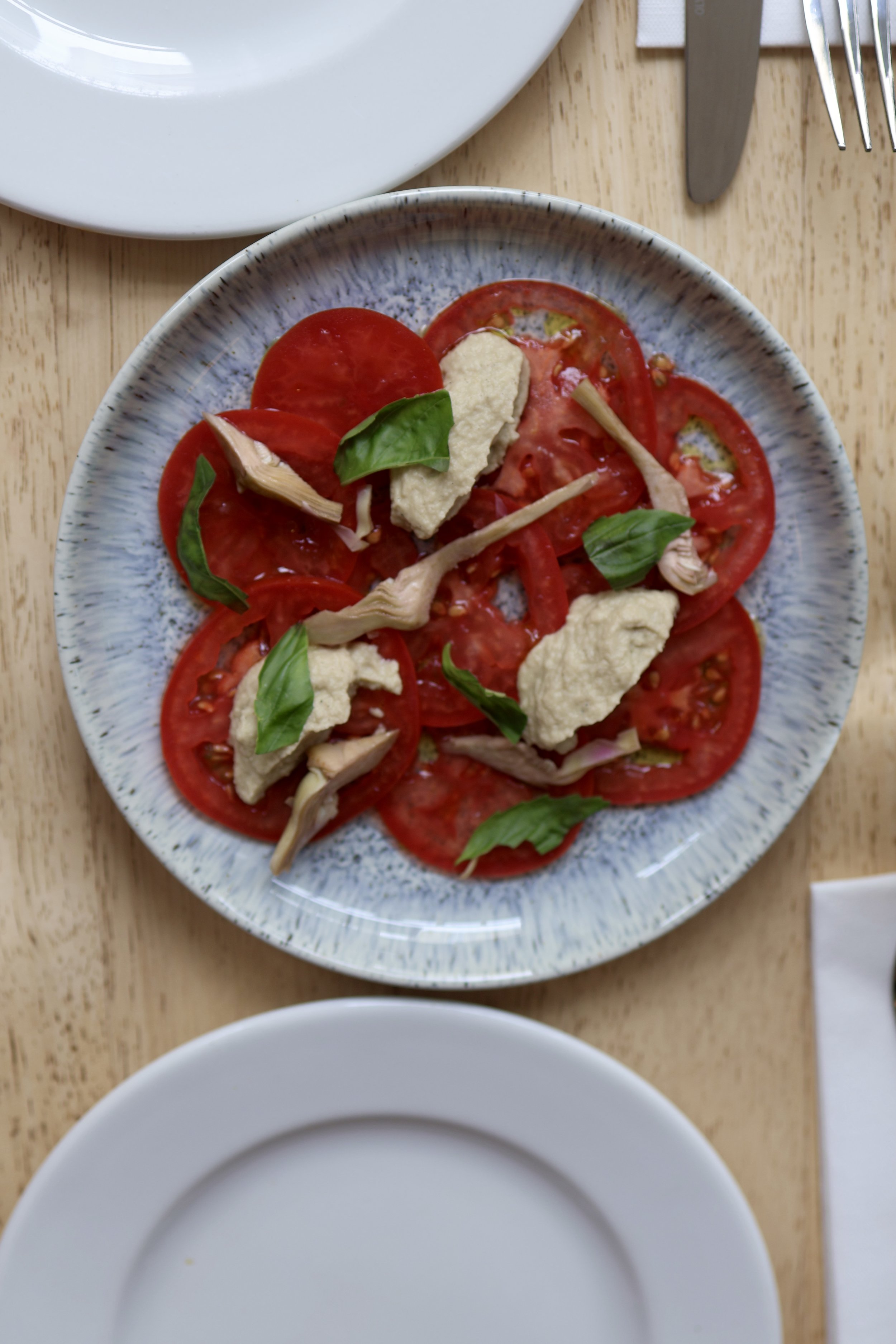 A plate with sliced tomatoes, mozzarella cheese, basil leaves, and artichoke hearts on a wooden table, with empty white plates nearby.