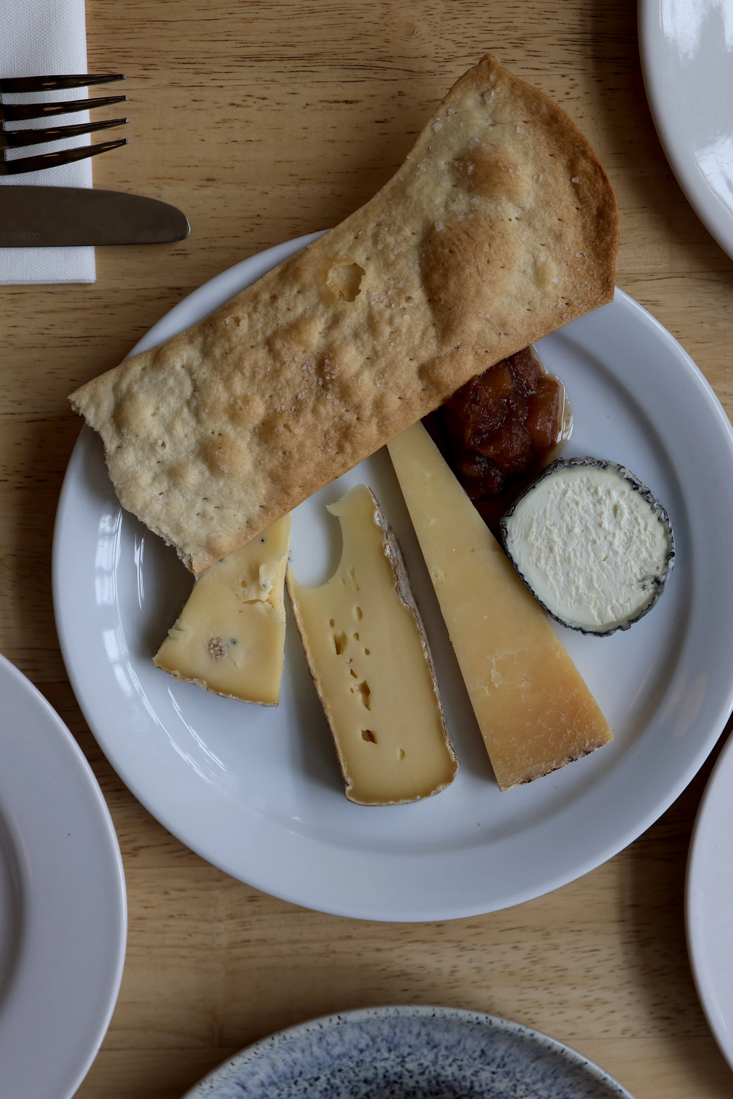 A white plate with a large piece of fried bread and several slices of cheese on a wooden table, with a small dollop of jam or chutney.