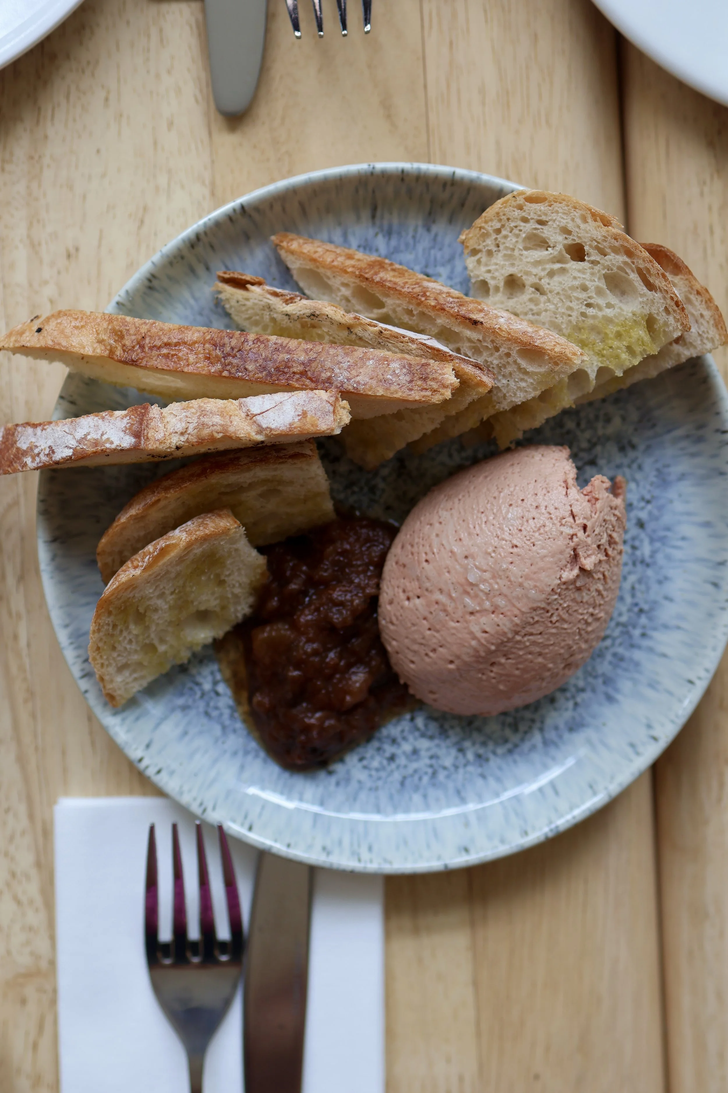 A plate of sliced bread with a side of jam, accompanied by a scoop of pink ice cream on a wooden table set with cutlery.