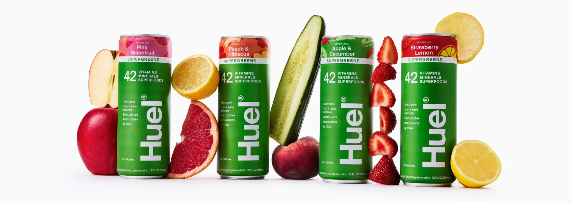 Four cans of Huel supergreens drink with different fruit flavors, surrounded by various fresh fruits including apples, lemon, grapefruit, peach, cucumber, strawberries, and lemon slices.
