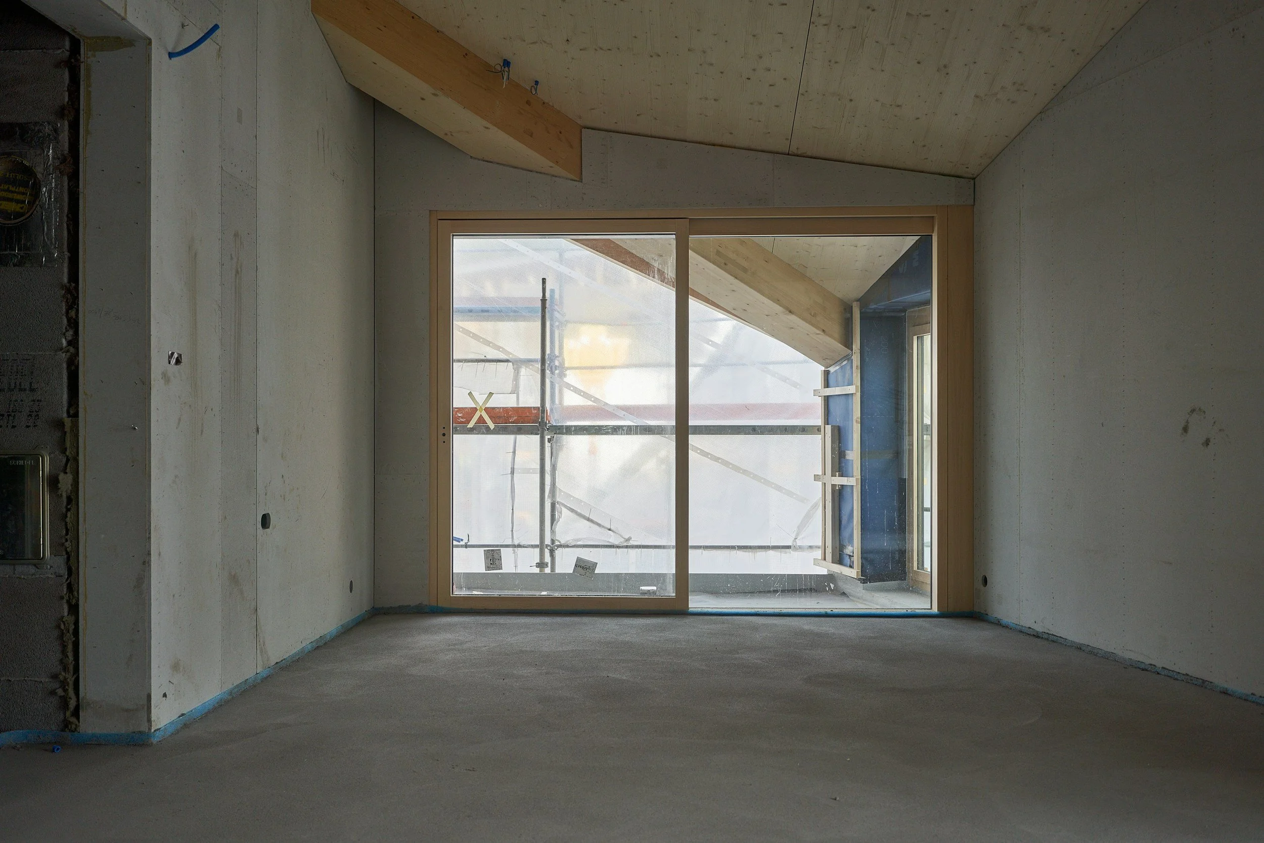 An unfinished interior room with concrete floors and walls, a large glass sliding door leading to an outdoor balcony, and wooden ceiling and trim, under construction.