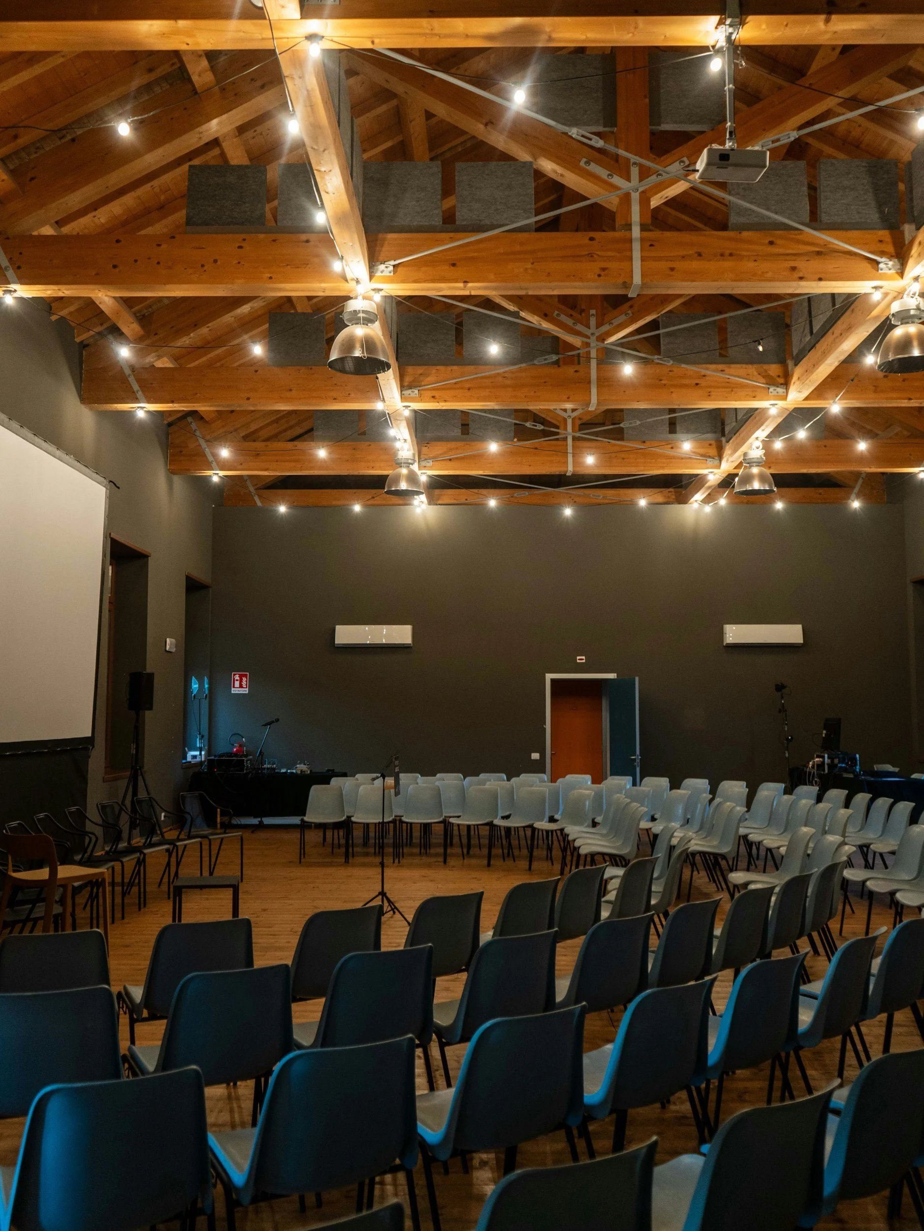 Indoor event space with rows of chairs arranged for a presentation or meeting, wood ceiling with exposed beams, string lights, and a stage area.