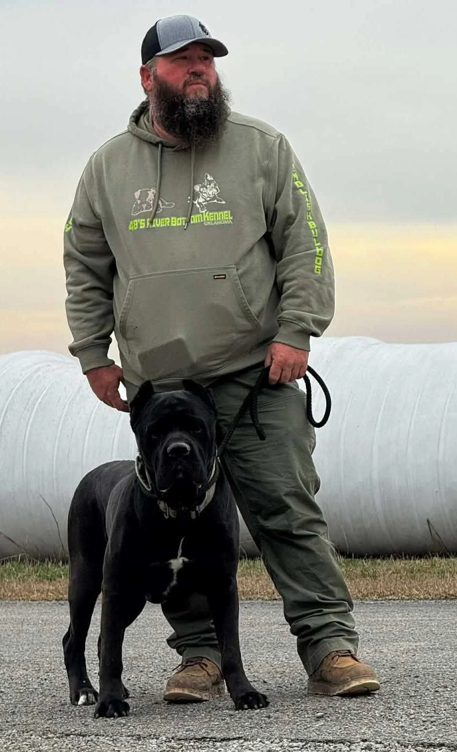 A man with a beard wearing a gray hoodie and a trucker hat stands outdoors holding a black dog on a leash.