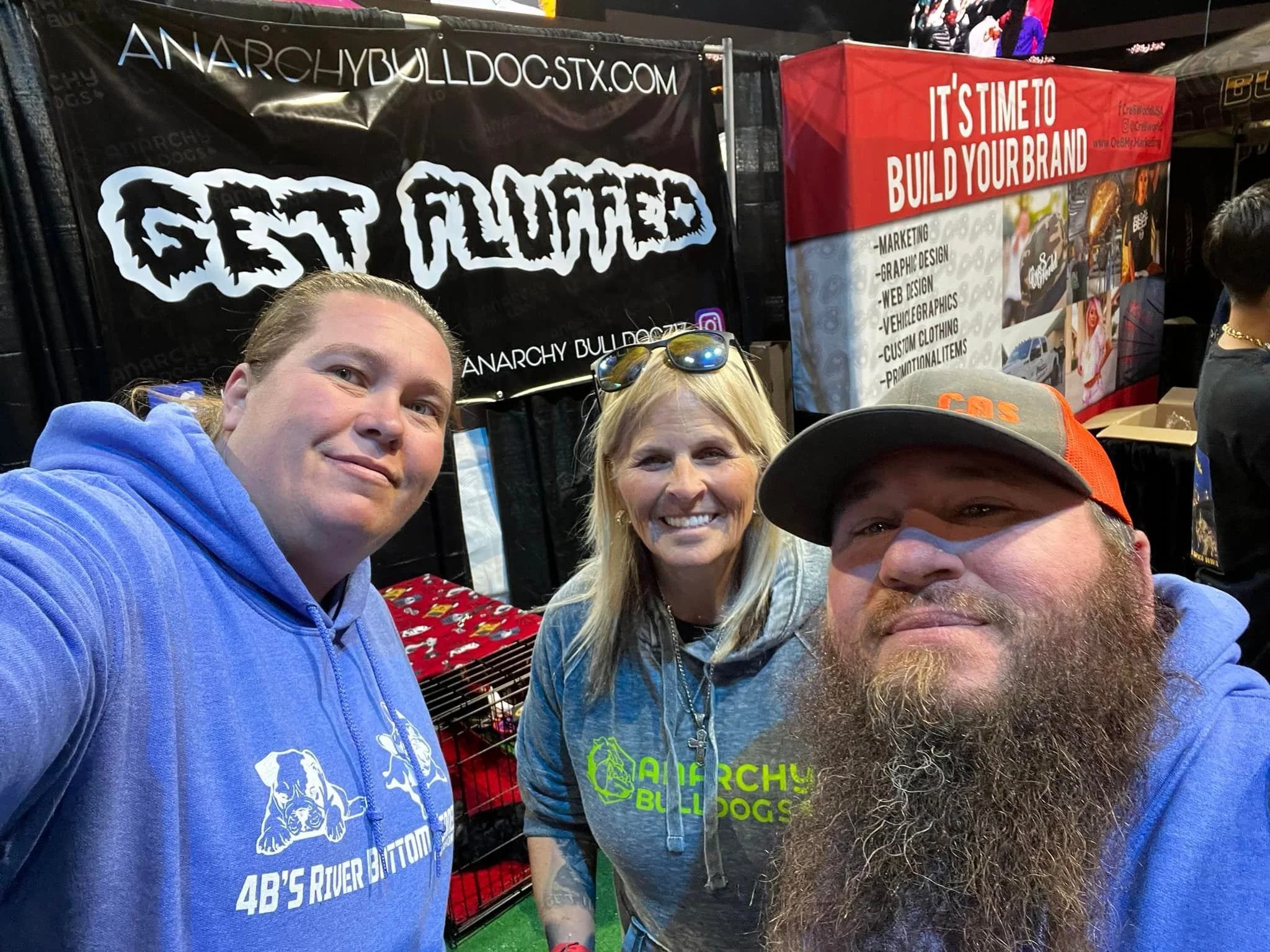 A group of people taking a selfie at a booth with banners advertising a website and services related to bulldogs. The banners indicate a focus on marketing, graphic design, web design, vehicle graphics, custom clothing, and promotional items. The people are smiling and wearing casual clothing, and the booth is decorated with various images and text related to bulldogs and branding.