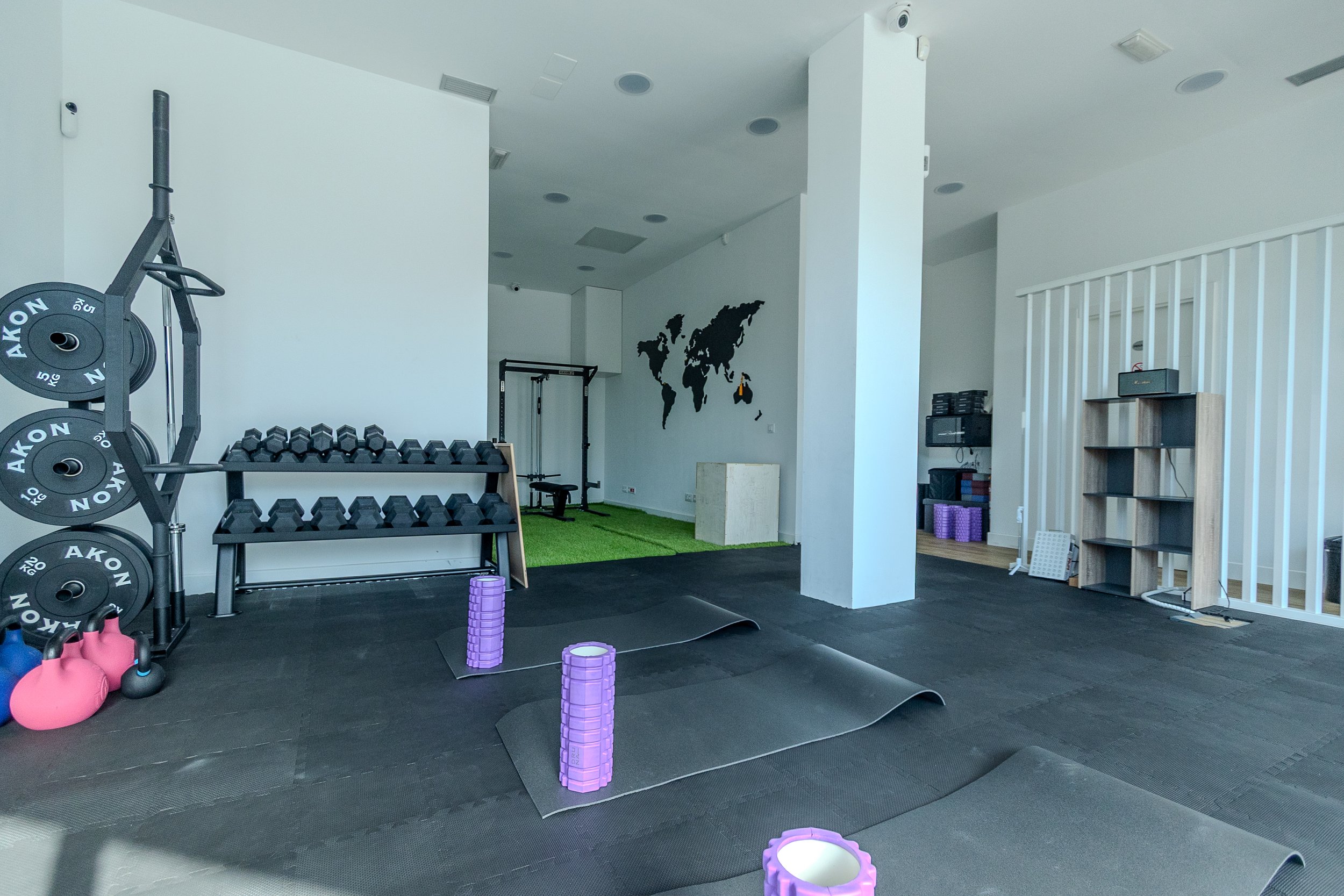 Espacio de ejercicio en un gimnasio con pesas, kettlebells rosas, esterillas, rodillos morados, y una pared con un mapa del mundo en negro.