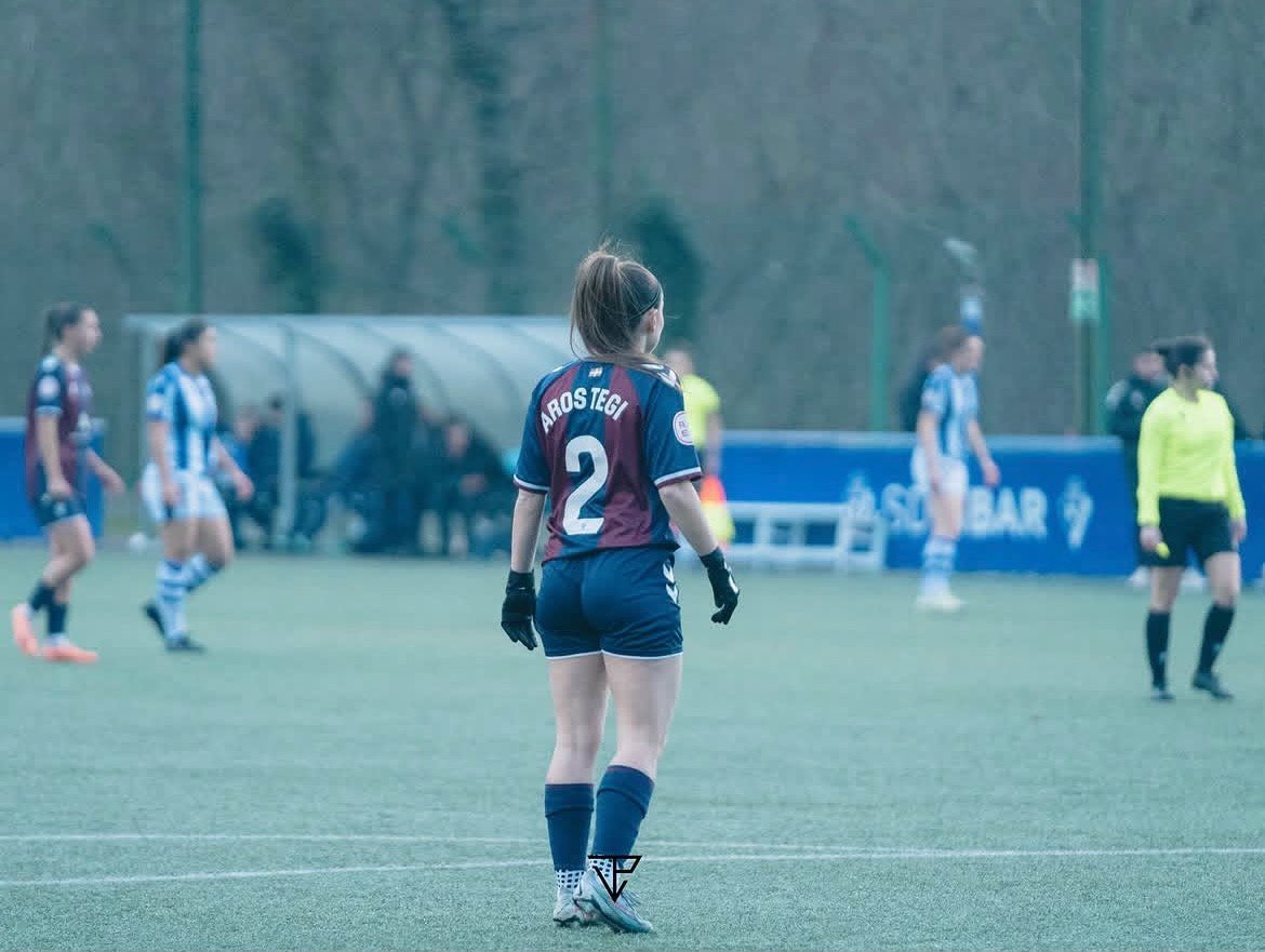 Jugador de fútbol femenino con uniforme azul y maroon, de espaldas, en un campo de fútbol durante un partido, con otras jugadoras y árbitra en el fondo.
