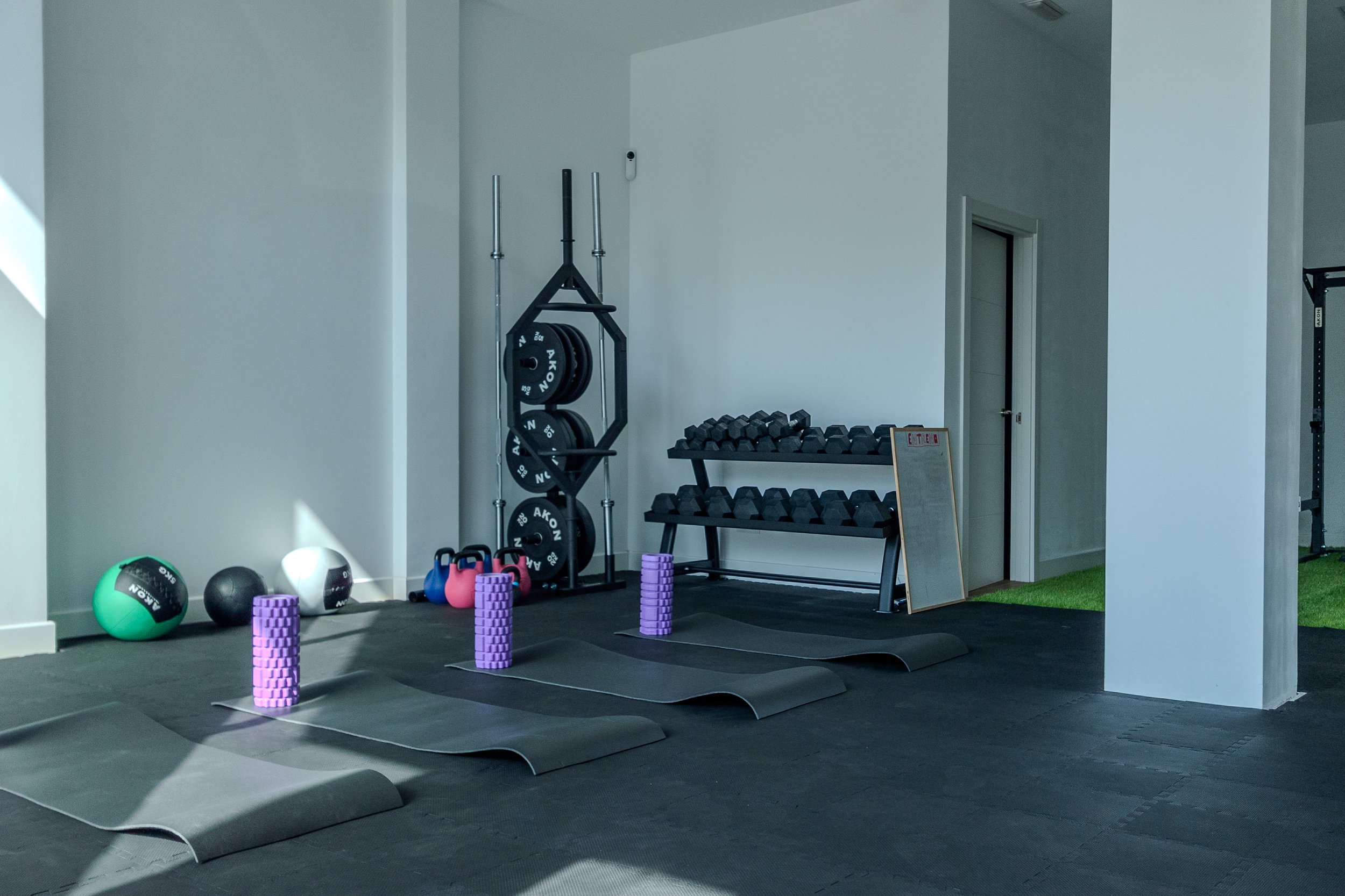 Espacio de gimnasio con pesas, balones de ejercicio, kettlebells, y esterillas en el suelo, con paredes blancas y una puerta al fondo