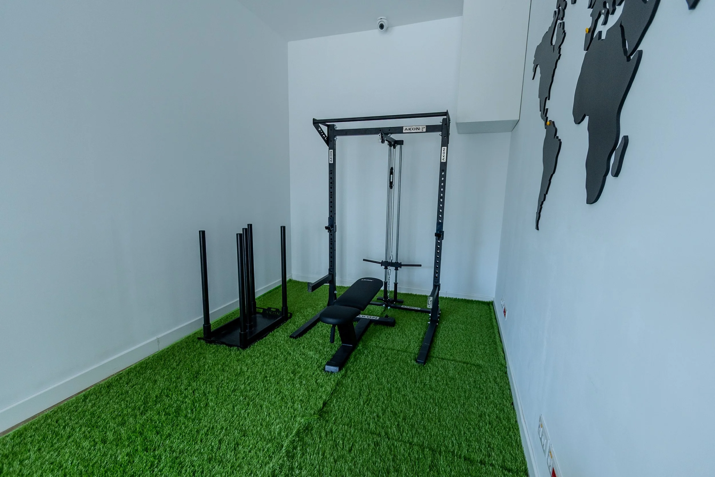 Gimnasio con paredes blancas, piso de alfombra verde y equipamiento de entrenamiento, incluido un marco para tracción y una banca de entrenamiento. También hay un mapa del mundo en la pared.