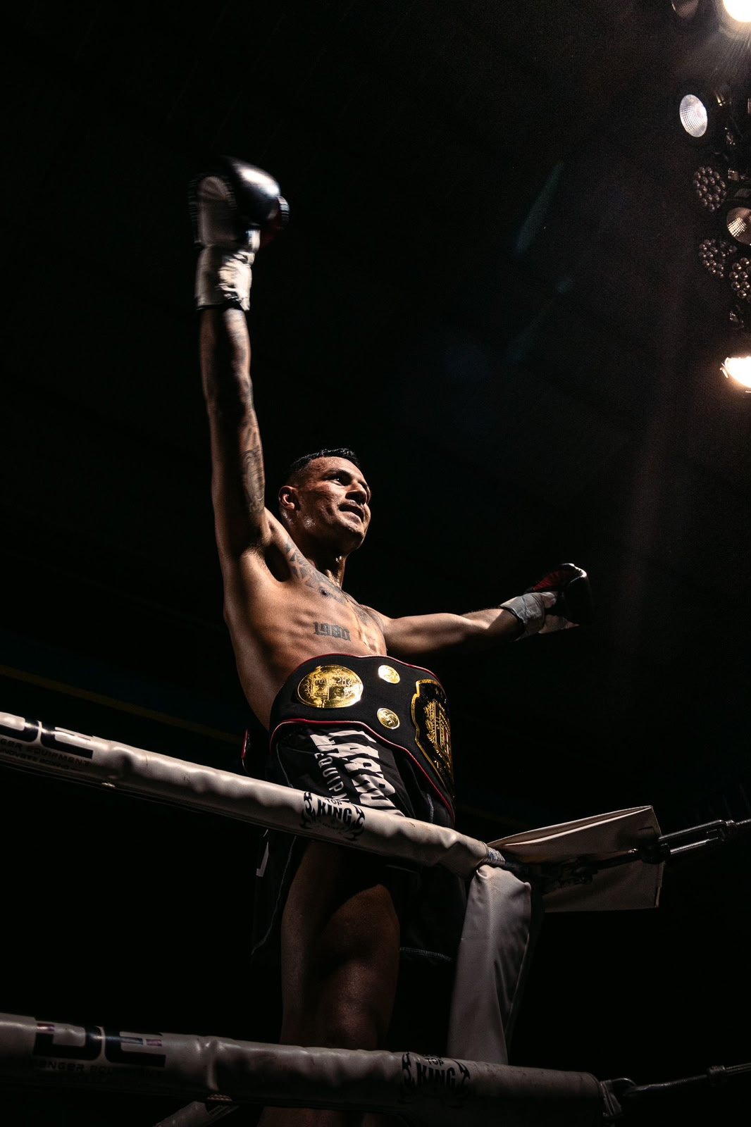 Campeón de boxeo con cinturón en el ring, levantando un brazo en señal de victoria, en un ambiente oscuro con luces.