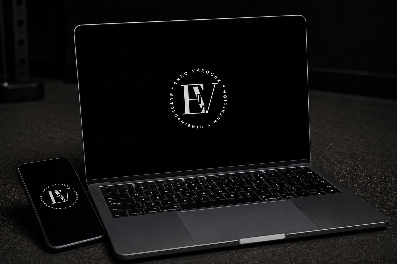 Una laptop y un teléfono móvil en un escritorio de color oscuro, ambos con el logo de Enrique Vázquez, Entrenamiento y Nutrición en la pantalla.