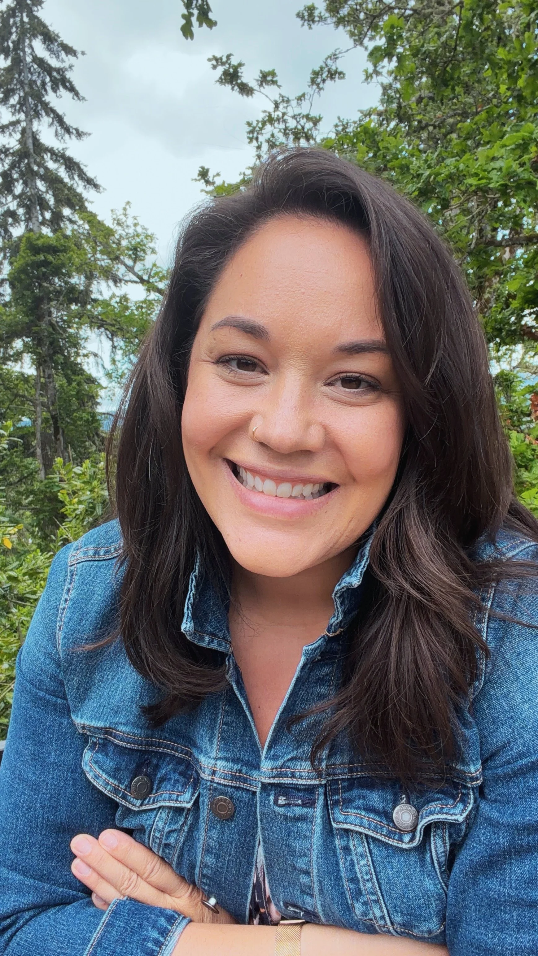 A woman with long dark hair smiling outdoors, wearing a blue denim jacket, standing in front of trees and cloudy sky.