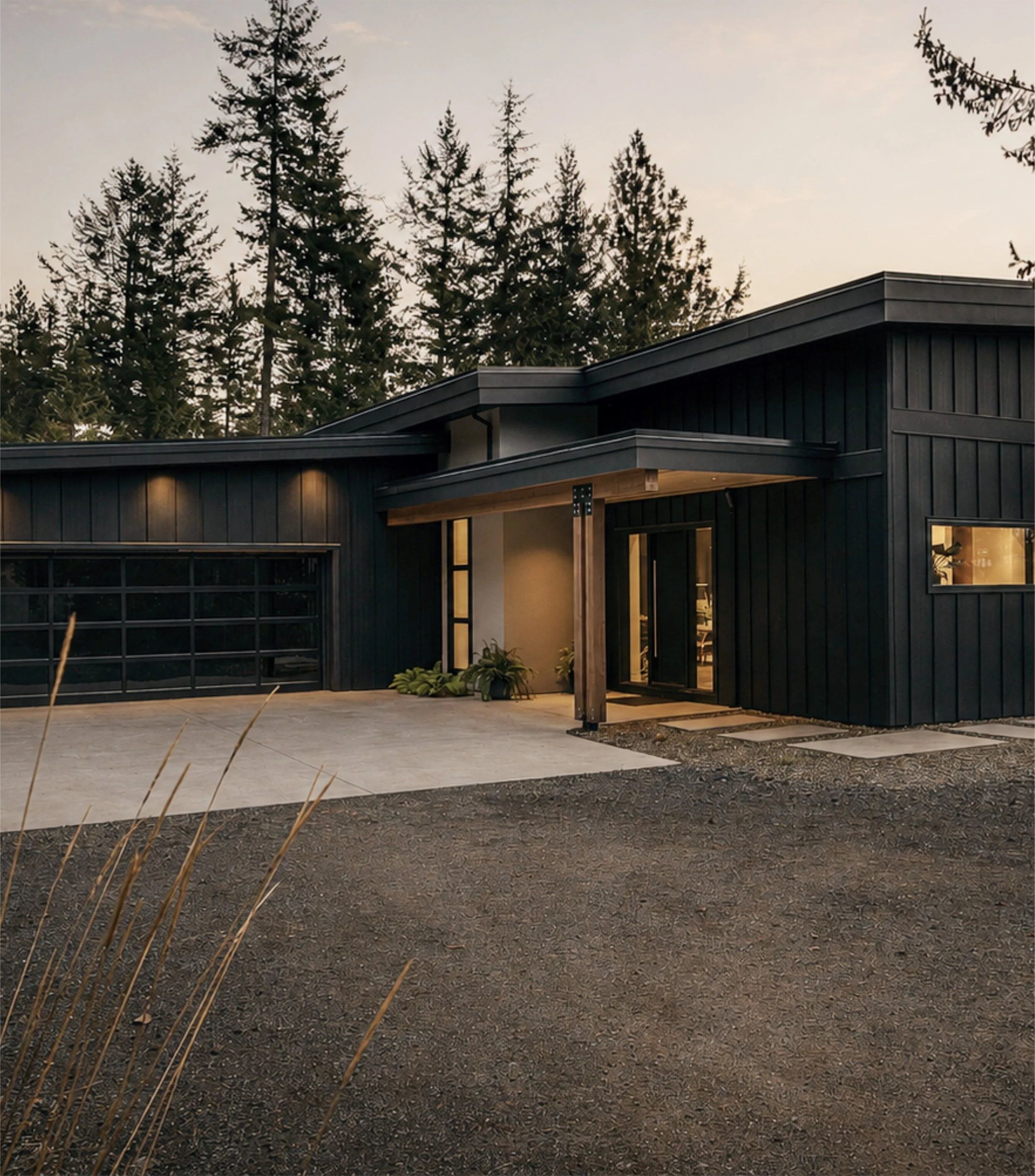 A modern house with black exterior walls, large windows, and a wood pillar, situated in a wooded area with tall pine trees visible in the background at dusk.