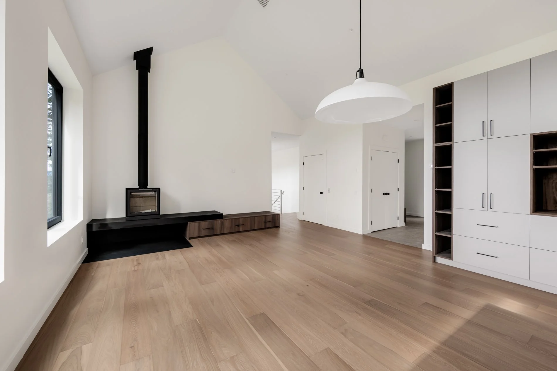 Interior of a modern, minimalistic living area with white walls, large window, wooden flooring, a wood-burning stove, and built-in white cabinets.