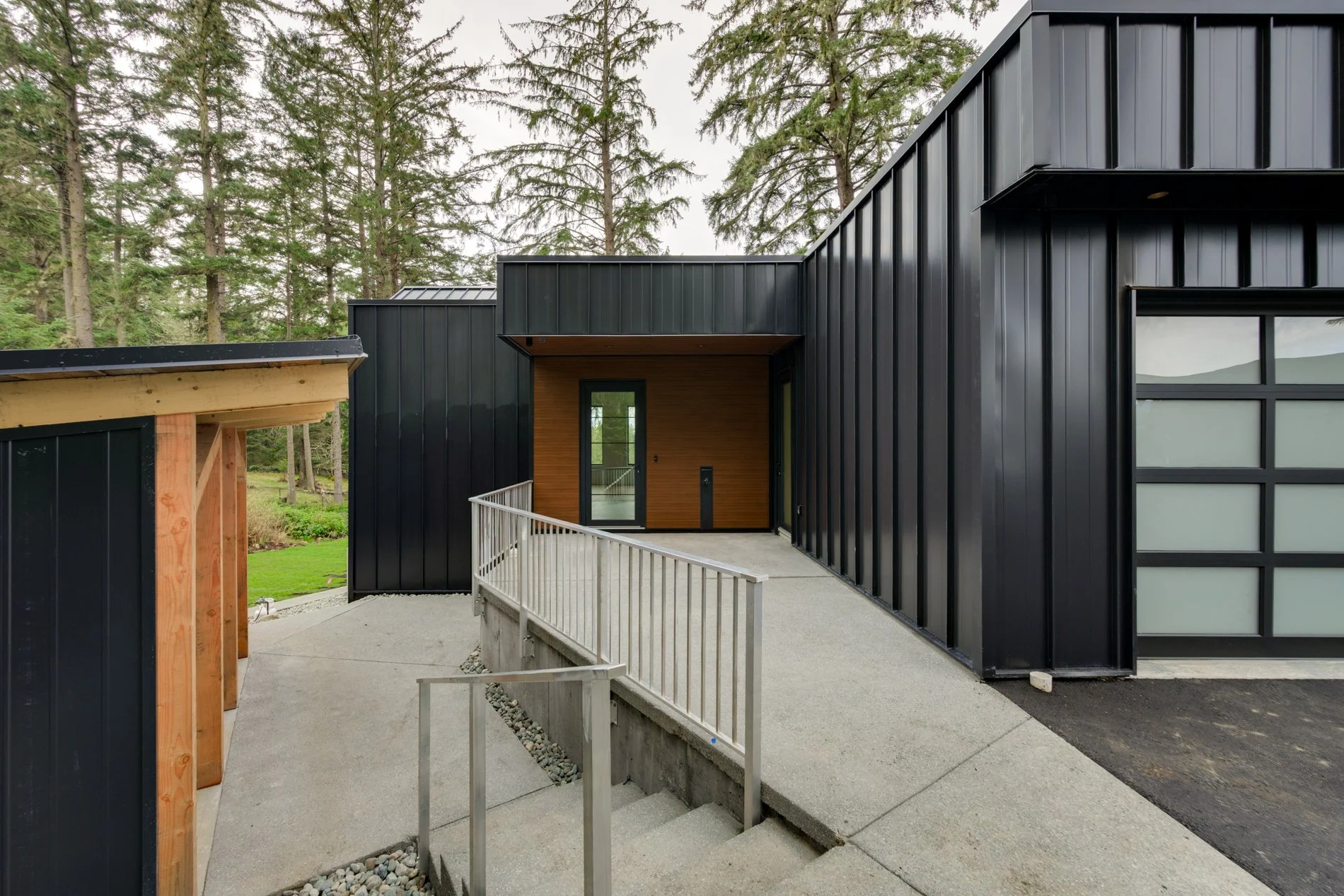 Modern black metal house with wood-paneled entrance, surrounded by pine trees, with a concrete pathway and metal railing leading to the door.
