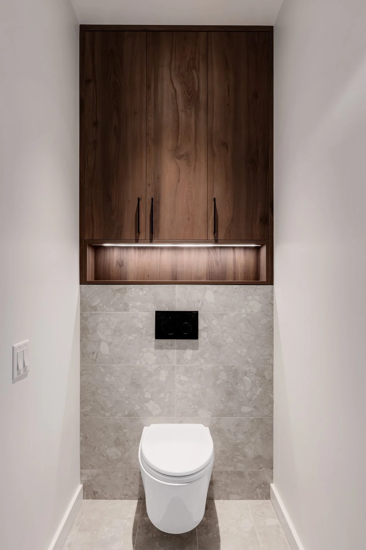 A modern, narrow bathroom with white walls, a wooden cabinet above the toilet, gray stone-like tiles on the wall and floor, a black flush plate above the toilet, and a dual light switch on the left wall.