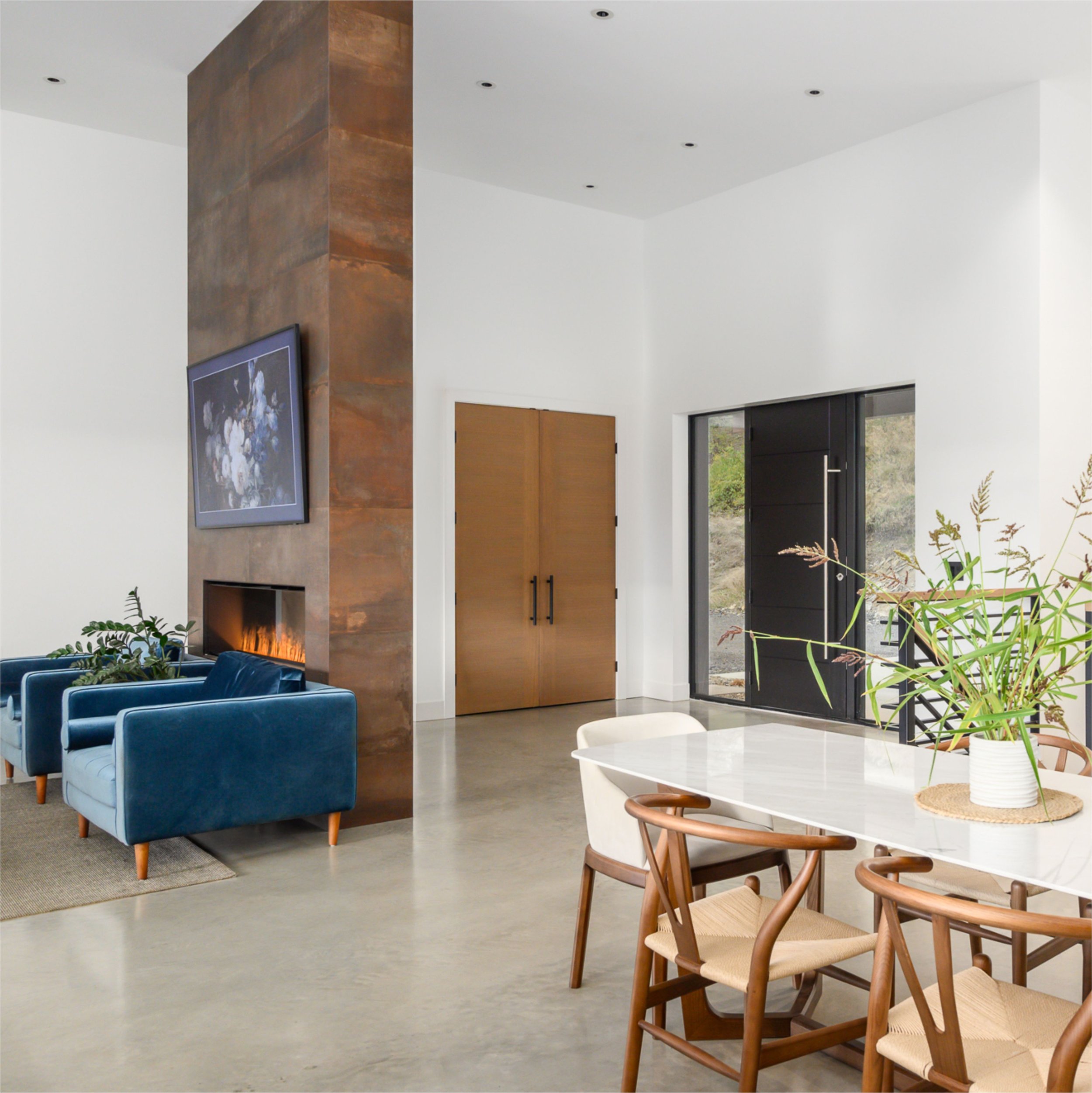 Modern interior living space with blue velvet sofas, wooden chairs with woven seats, a white marble table, and a textured rust-colored wall with a mounted television. Large windows showcase an outdoor scene, and there are plants near the windows.