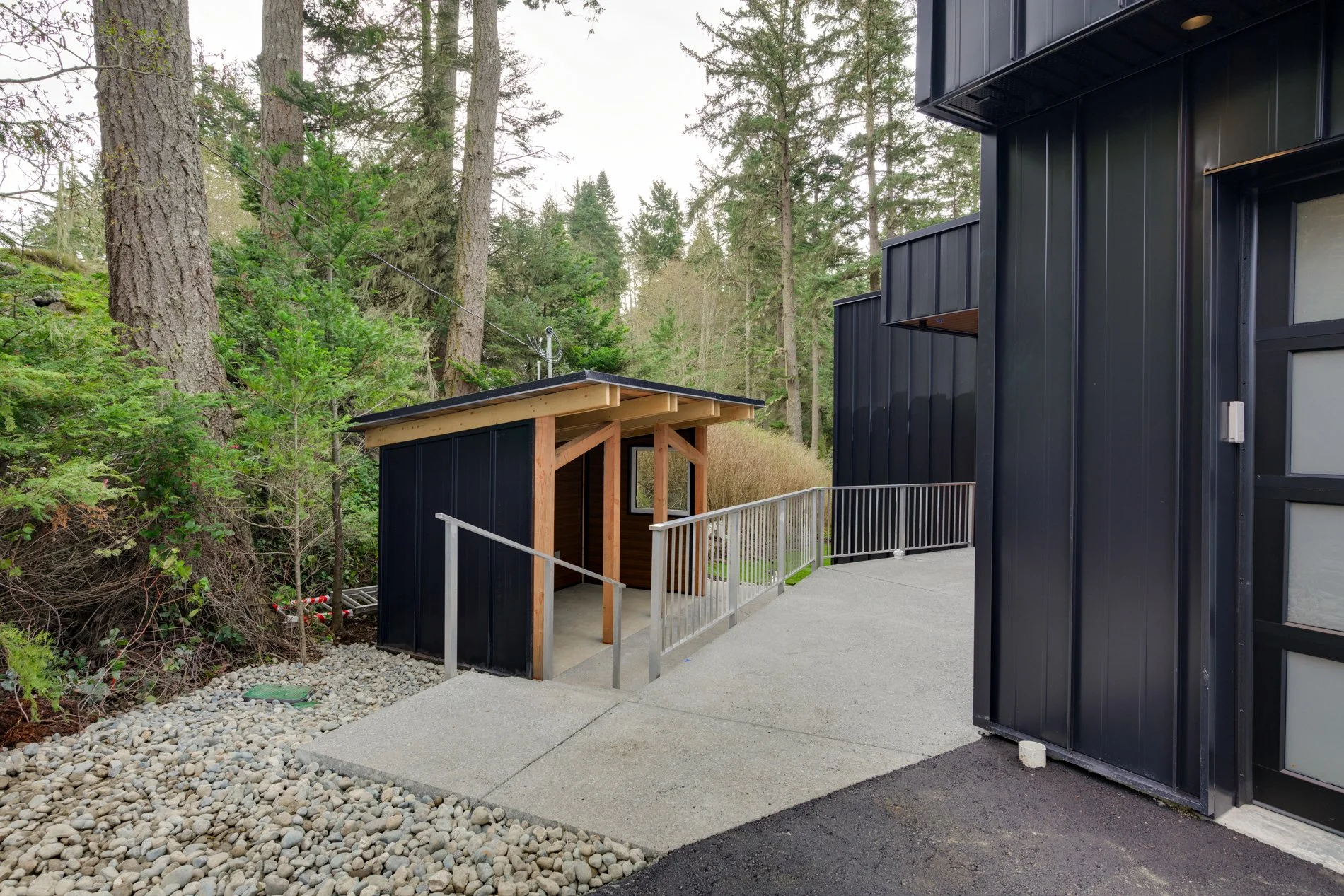 Modern black building exterior with metal siding, window, and a small wooden covered entryway. Surrounding natural landscape with tall trees and dense forest background. Concrete and gravel walkways.