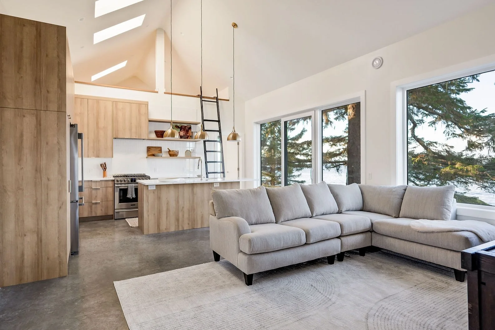 Open-concept living room and kitchen with large windows showing a view of trees and water, beige L-shaped sofa, wood cabinets, and pendant lights.