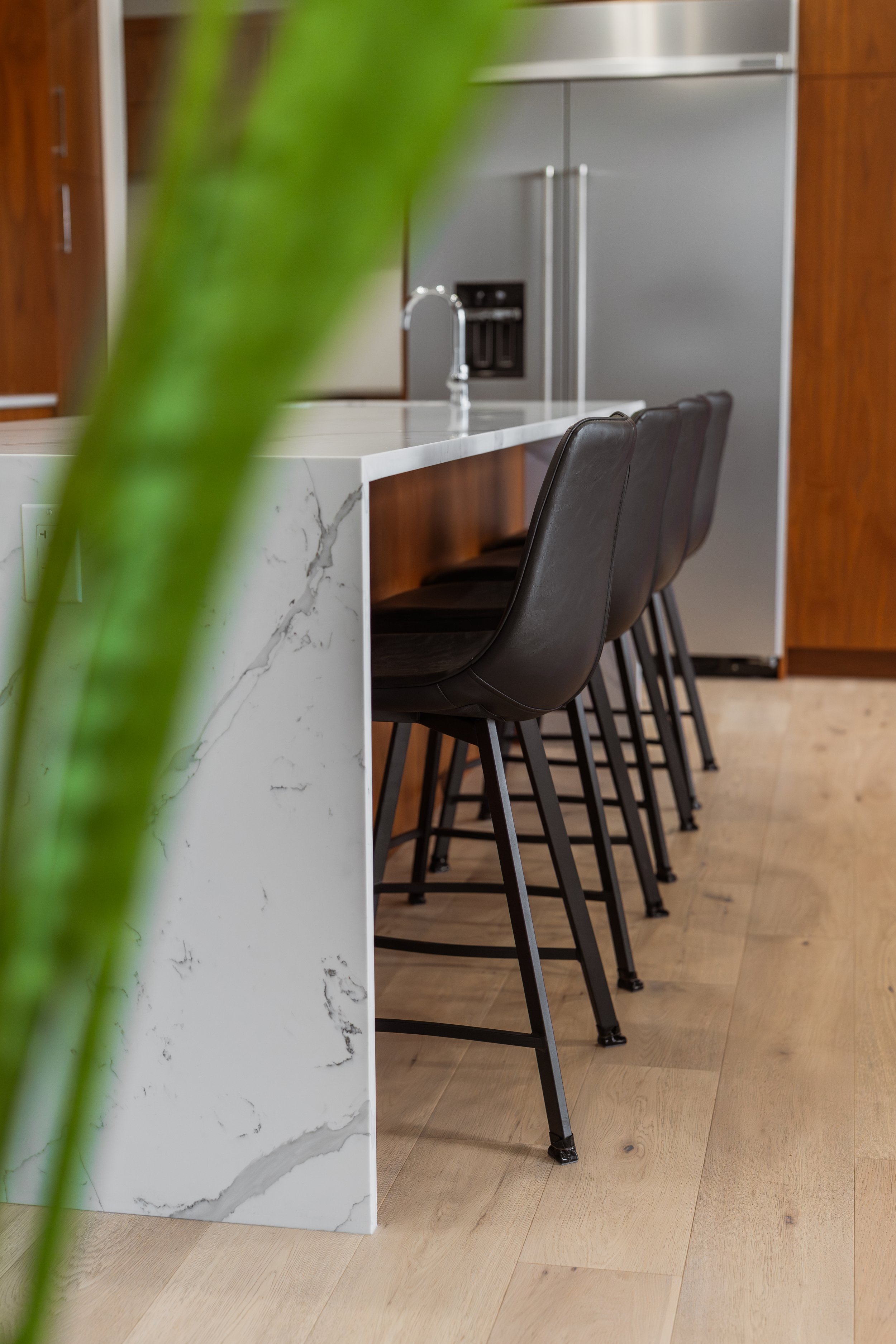 Modern kitchen with a white marble island, black bar stools, a stainless steel refrigerator, and wooden cabinets.