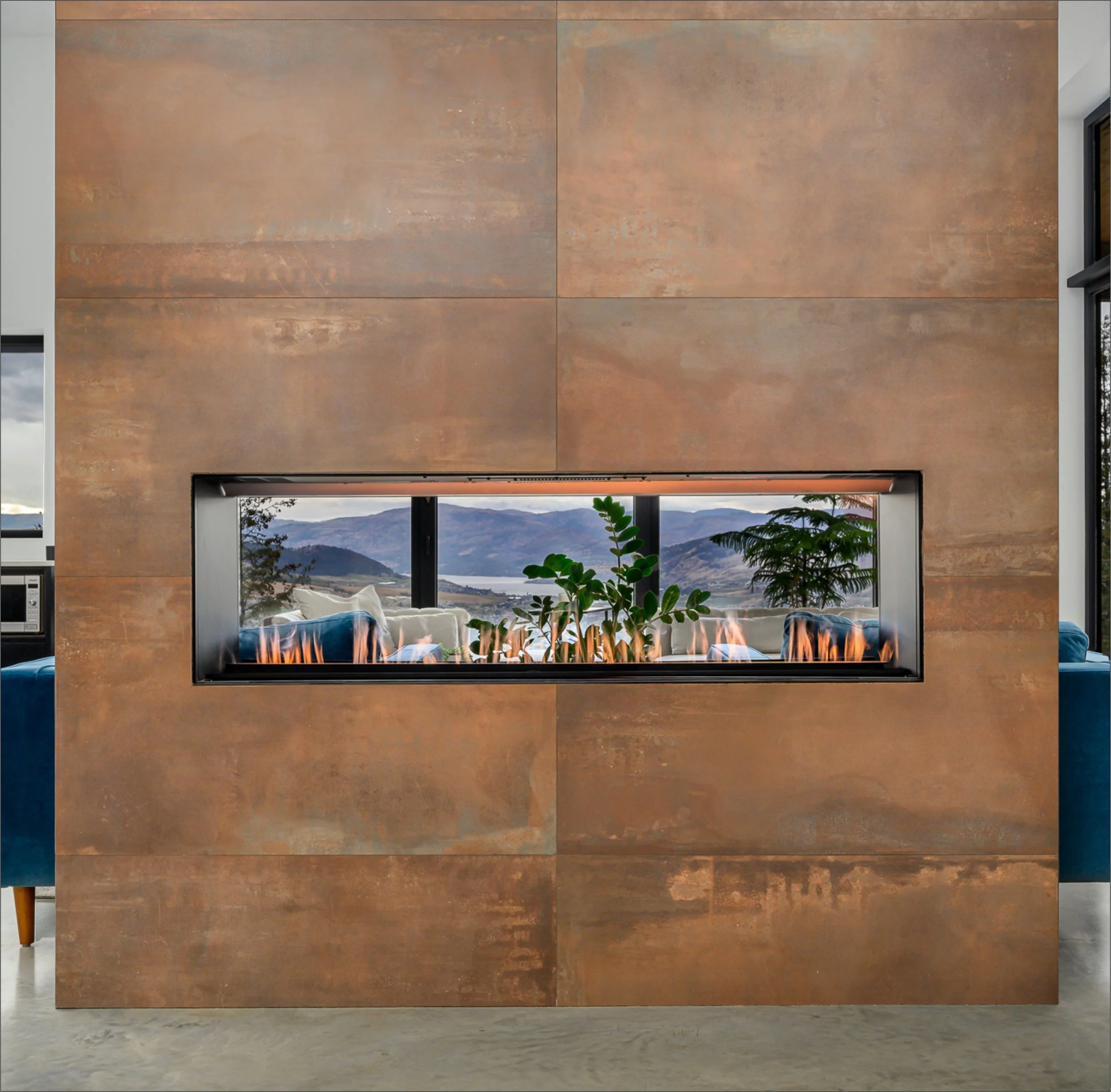 Interior view of a modern house with a fireplace, large window overlooking mountains and lake, and leafy green plants.