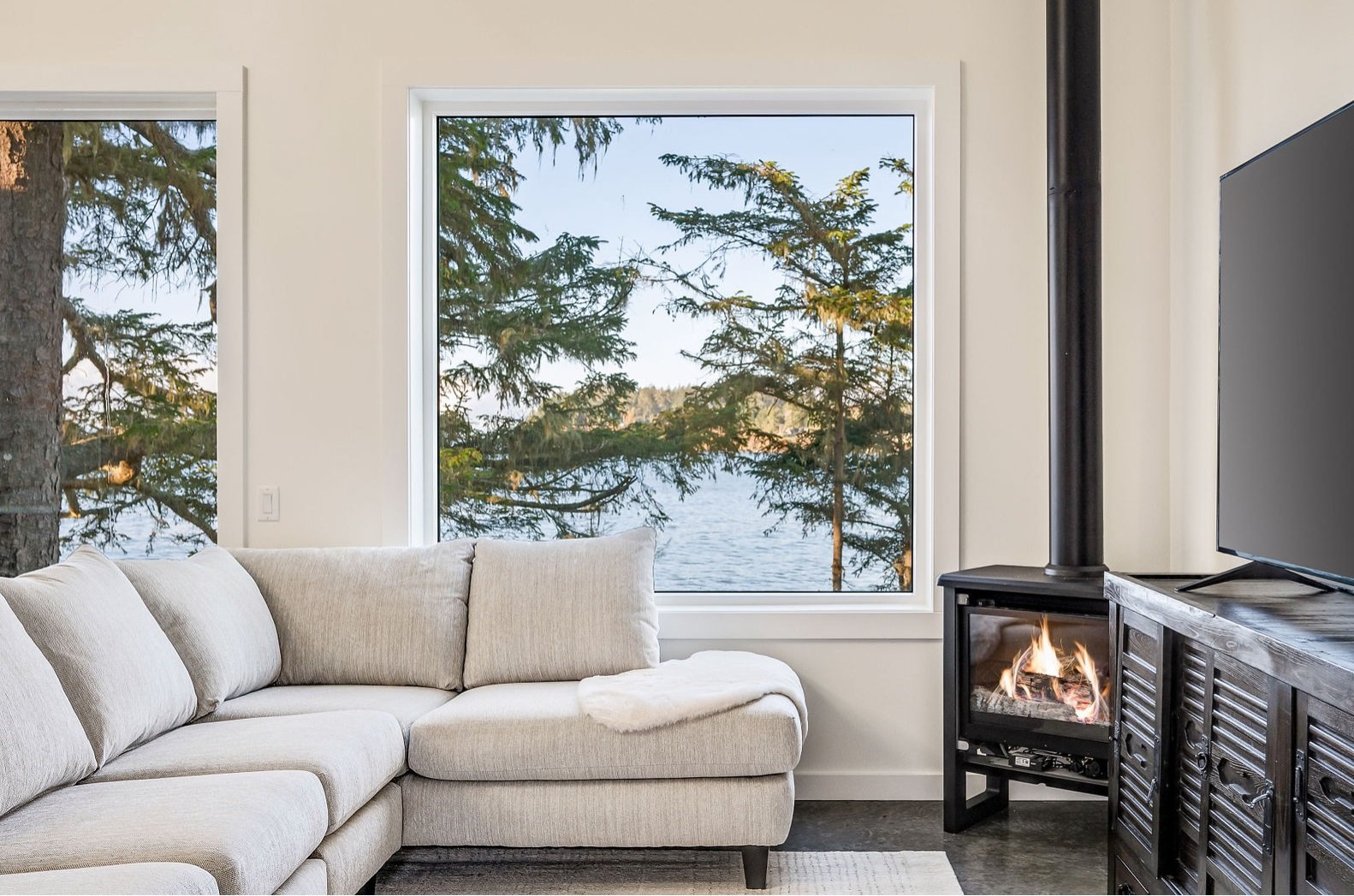 Living room with beige sectional sofa, large window showing trees and lake, and a wood-burning stove with flames.