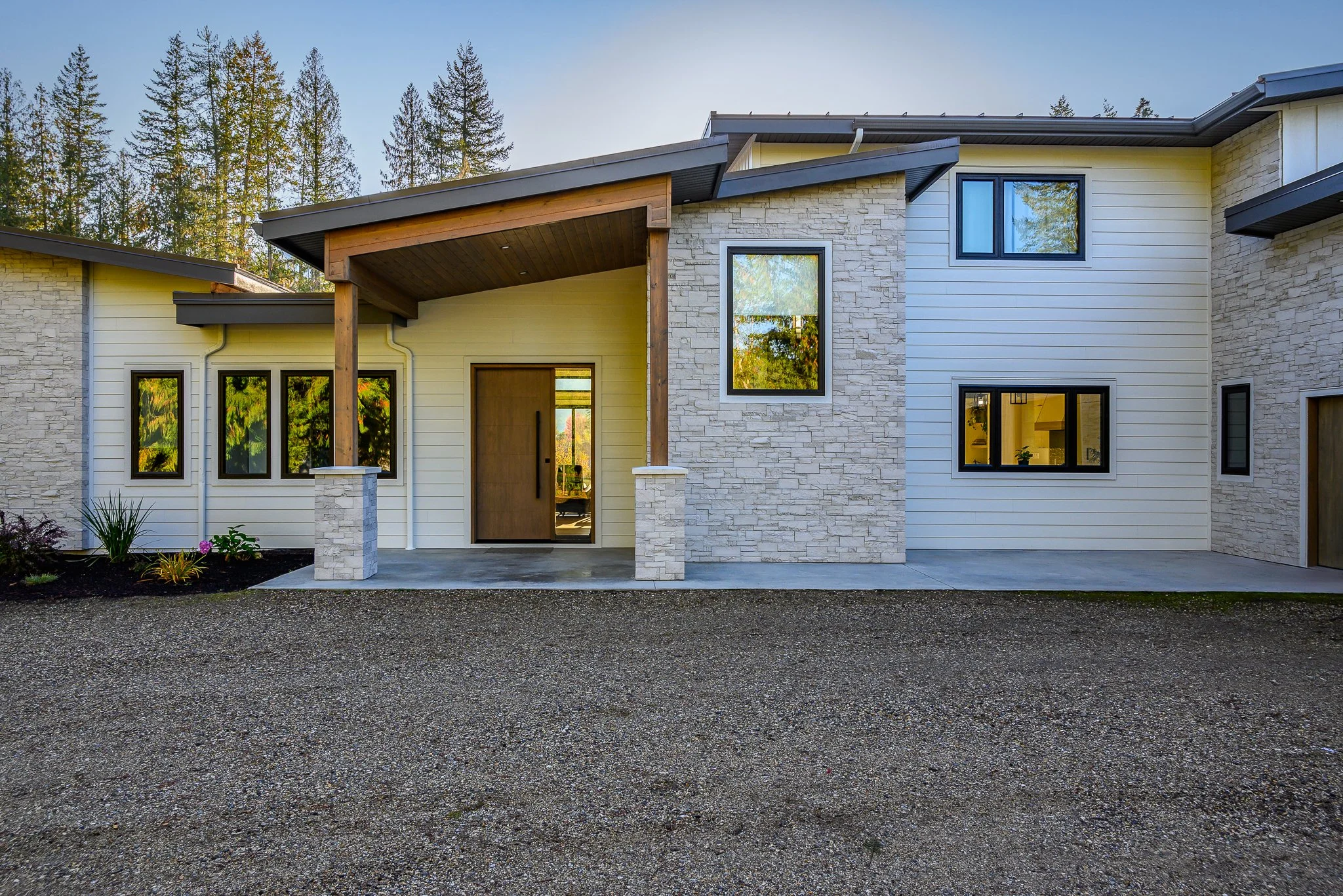 Front view of a modern house with a stone and siding exterior, multiple windows, and a wooden covered entrance, surrounded by tall trees and a gravel driveway.