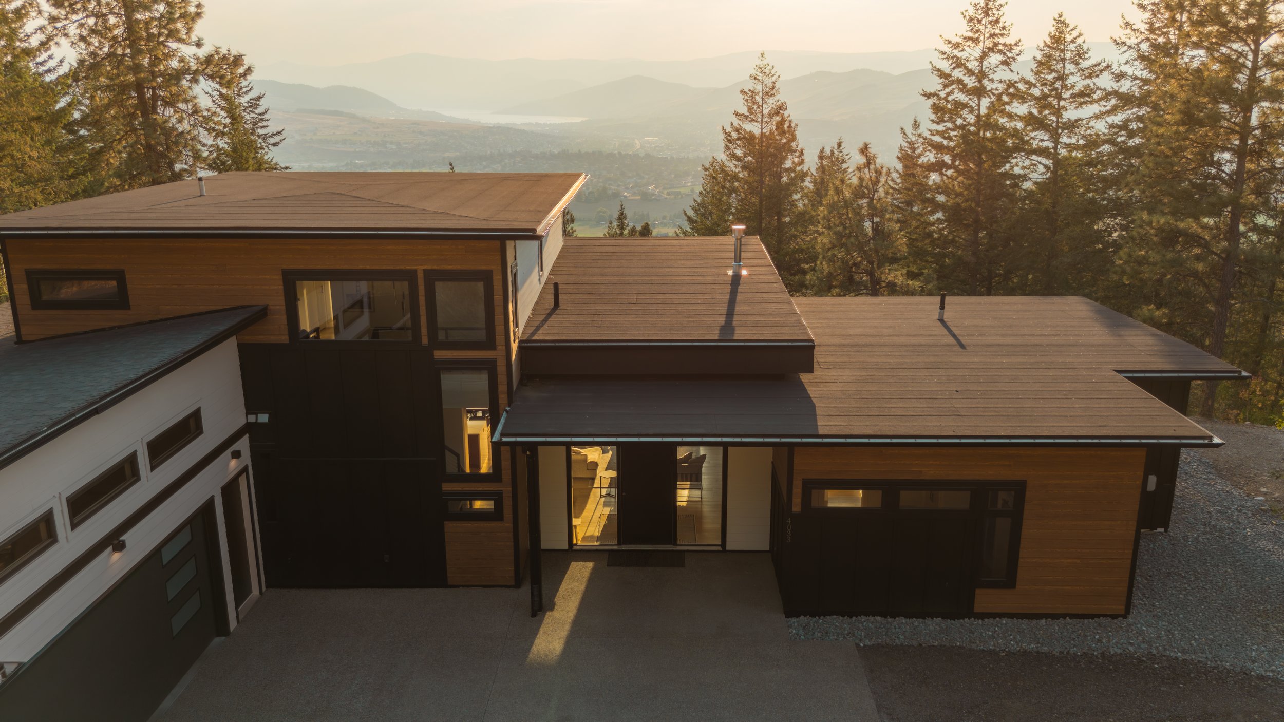 A modern house with flat roofs, large glass windows, and a mix of wood and black exterior walls, set against a backdrop of forested mountains and distant lake.