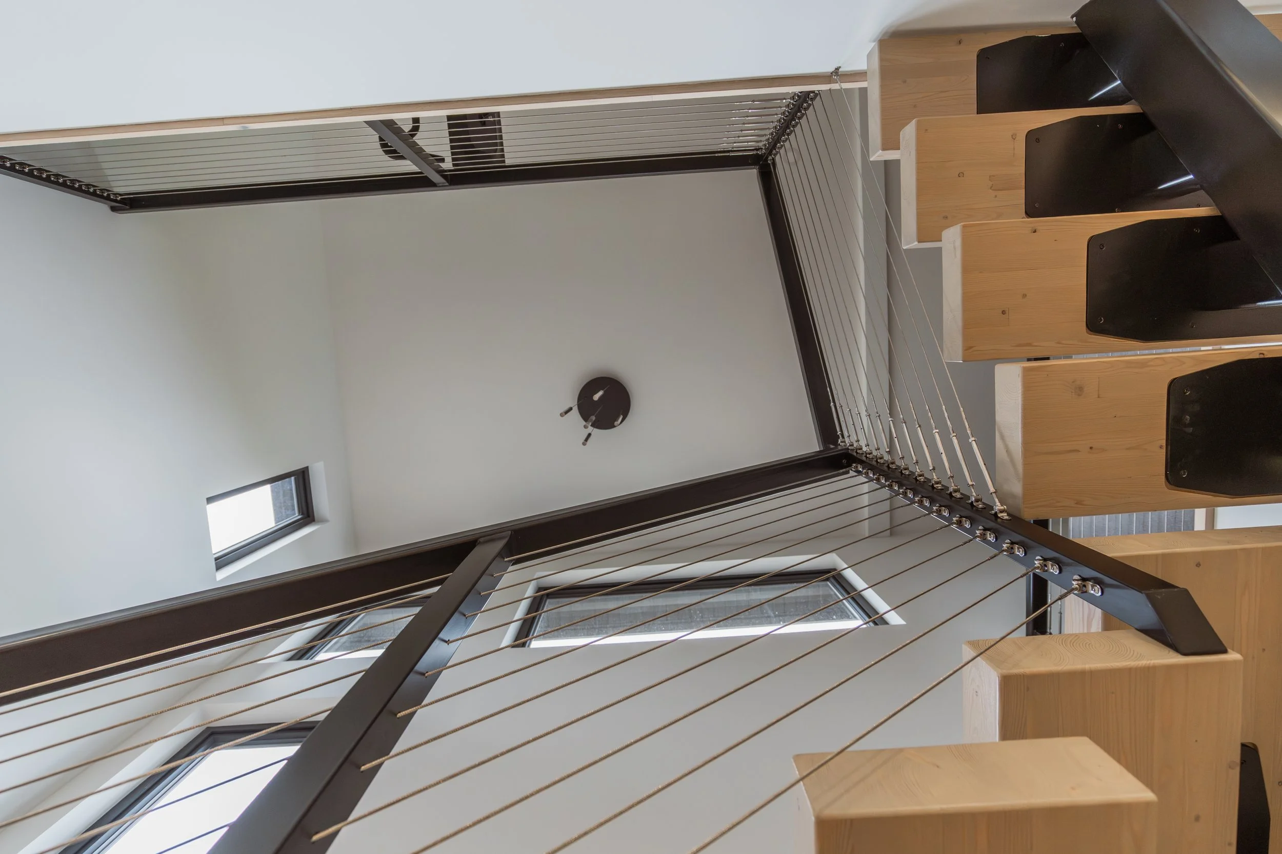Interior of a modern home with a staircase made of wooden steps and black metal railings, a wall-mounted clock, small windows, and a wire drying rack in the corner.