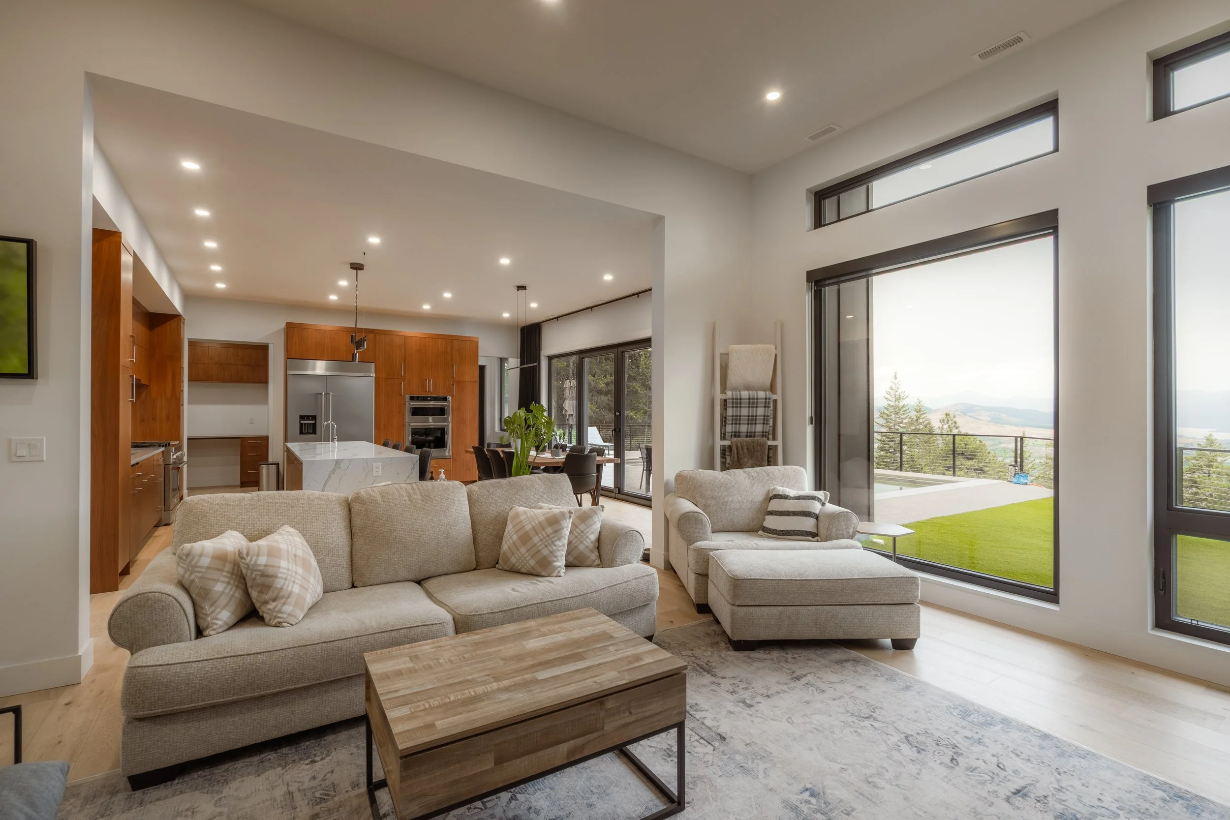 Open-concept living room and kitchen area with large windows showing a scenic outdoor landscape. The living room has a beige sofa with plaid pillows, a matching armchair, and a wooden coffee table. The kitchen features wooden cabinetry, a marble-topp