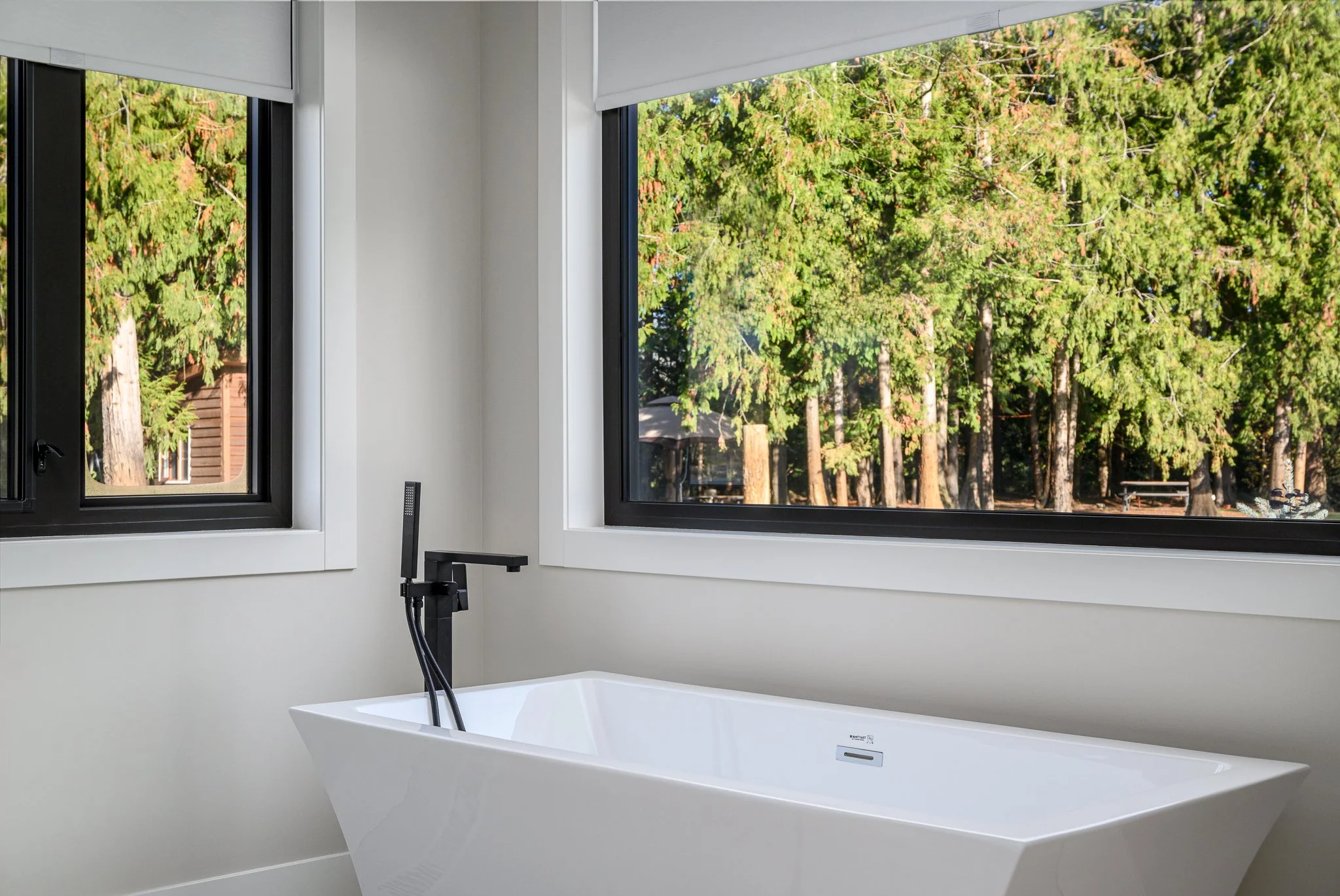 View from a bathroom with a window showing green trees outside, a white bathtub with a black faucet, and a small black object on the ledge.