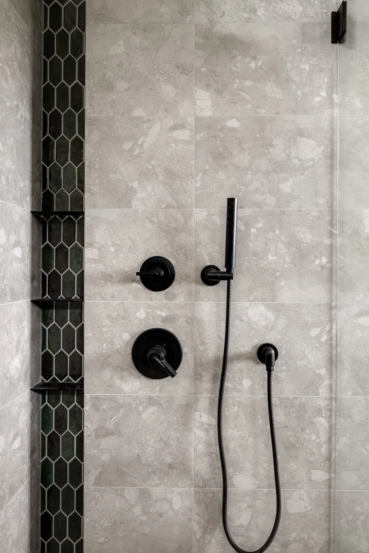 A modern shower with a black handheld showerhead, black control knobs, and a vertical black strip of hexagonal tiles on the left side. The wall is covered in light-colored marble tiles.