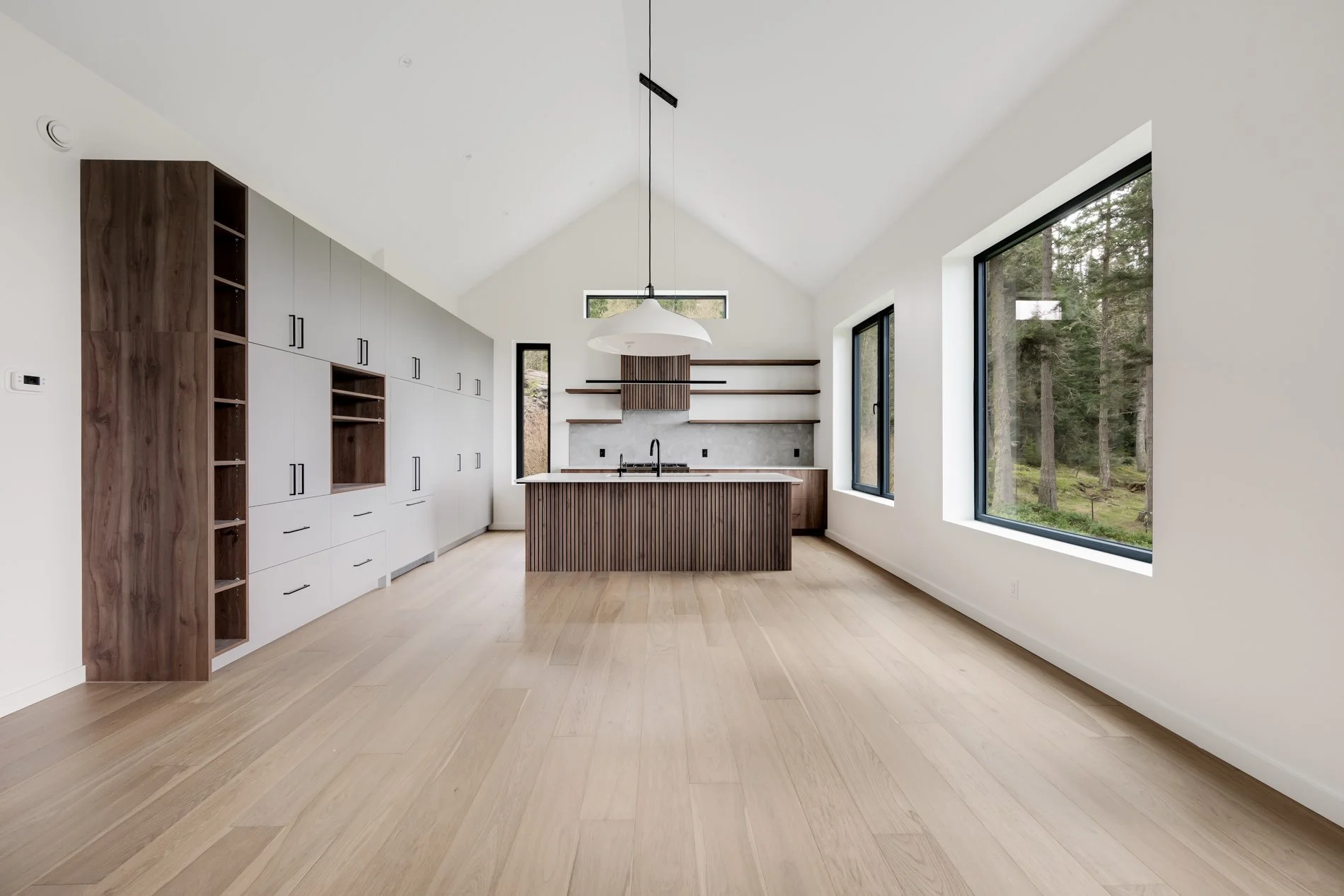 A modern kitchen with white walls, large windows showing trees outside, and wooden flooring. There are built-in cabinets and open shelving, with a central island featuring a dark wood finish and a white pendant light above.