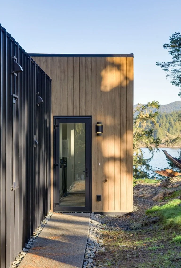 Modern house with black and wooden exterior, glass door, and a scenic outdoor view of trees, water, and mountains in the background.