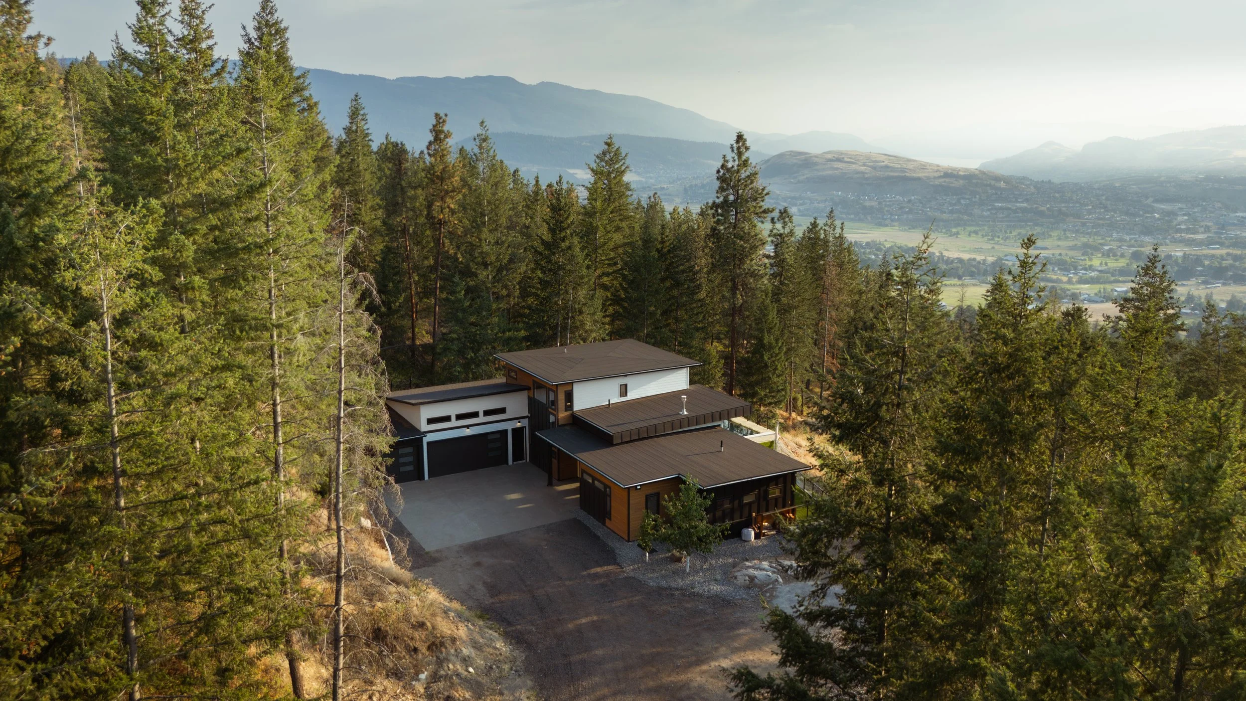Modern house with flat roofs amid tall evergreen trees on a hillside, overlooking a valley with distant mountains and a body of water.