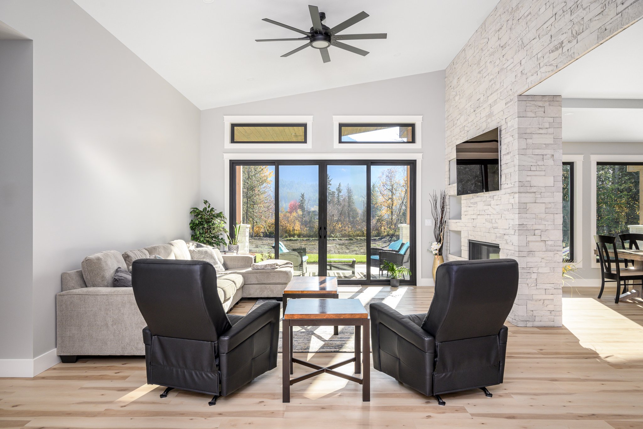 Modern living room with white walls, light wood flooring, and a white brick fireplace. Features a beige sofa, black armchairs, and a wooden coffee table. Large sliding glass doors open to a backyard with trees and outdoor seating.