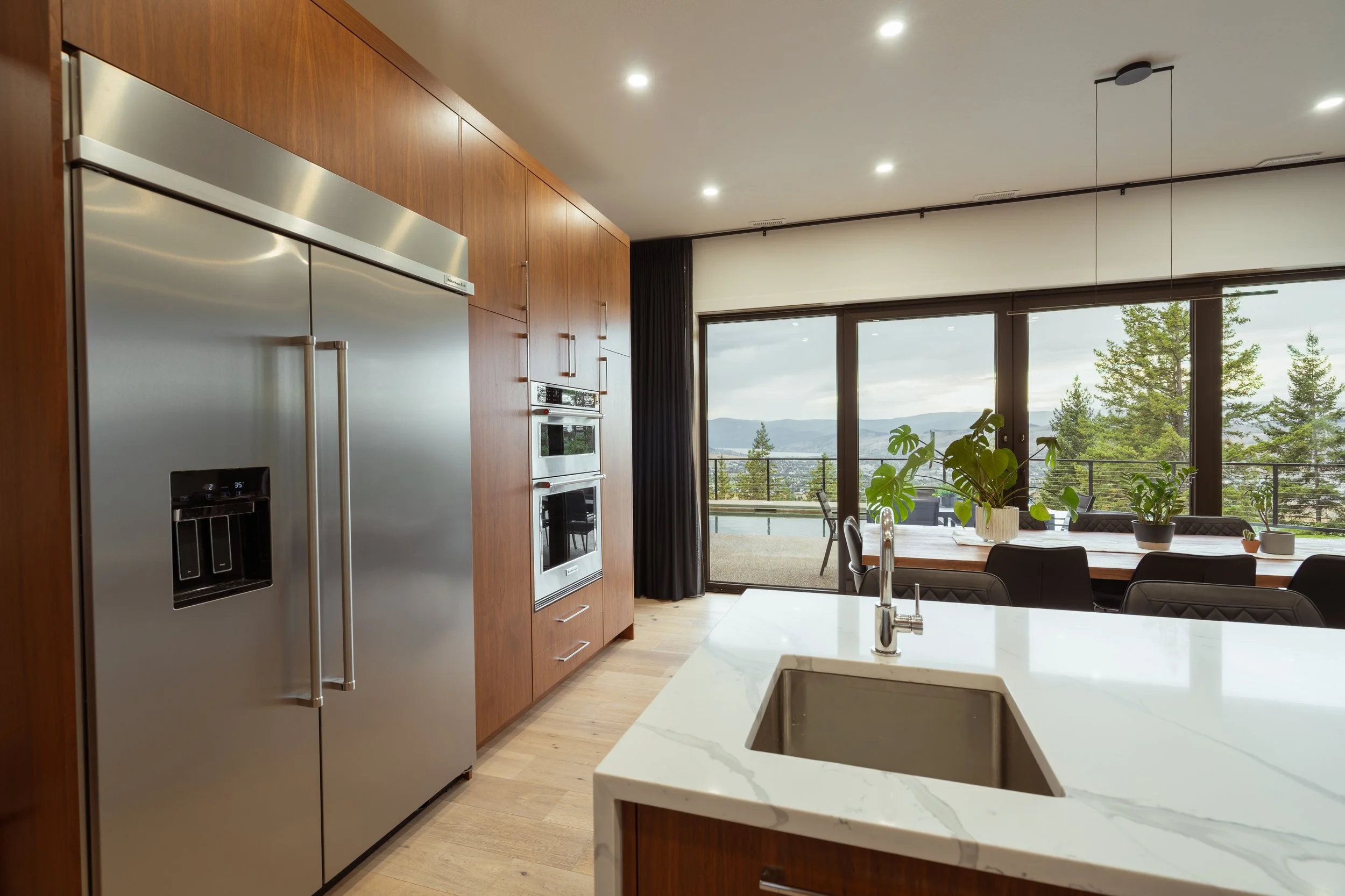Modern kitchen with stainless steel refrigerator, built-in oven, wooden cabinets, white marble island, large glass sliding doors opening to a balcony with mountain view, and indoor plants.