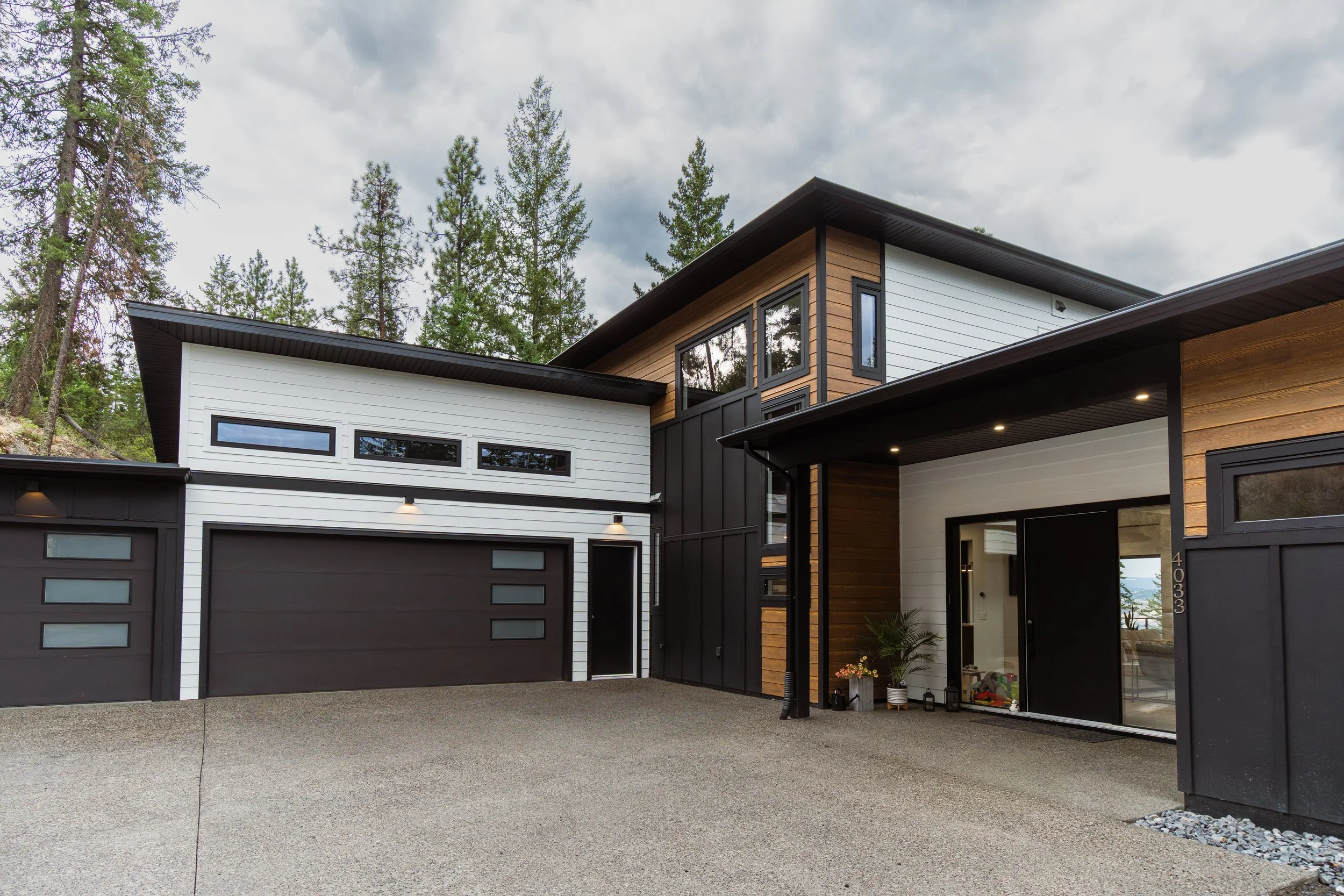 Modern house with black and white exterior, wood accents, large windows, and a driveway, surrounded by tall pine trees on a cloudy day.