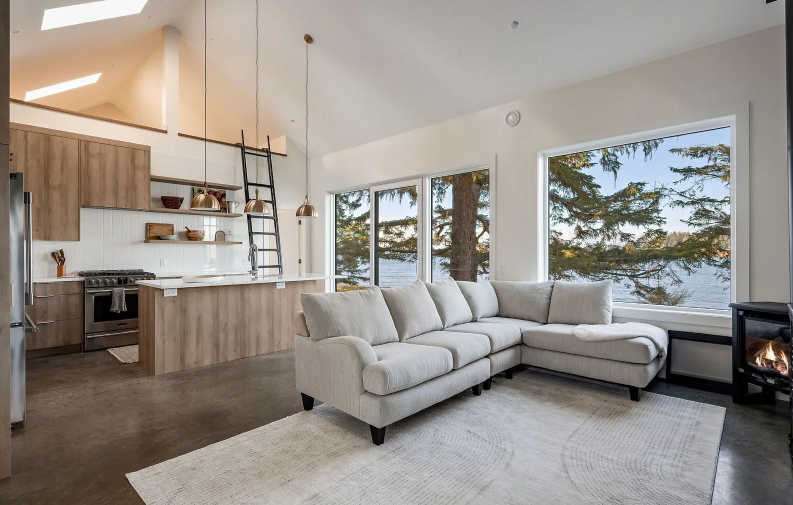 Open concept living room and kitchen with large windows showing a lake and trees outdoor. The living room has a light-colored sectional sofa, a rug, and a small electric fireplace. The kitchen features wooden cabinets, a white countertop, open shelvi